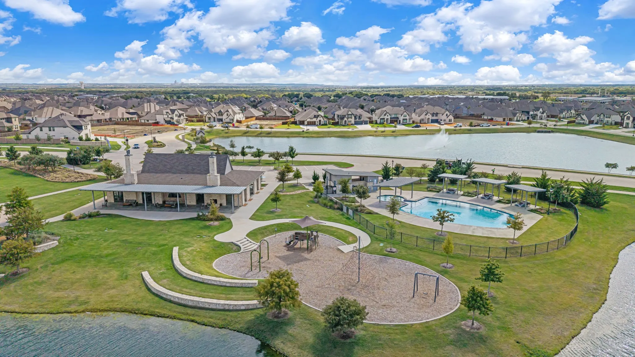 Community recreational complex featuring a swimming pool with pergola shade structures, a fenced playground area, stone retaining walls, and a multi-purpose pavilion situated on a waterfront lot