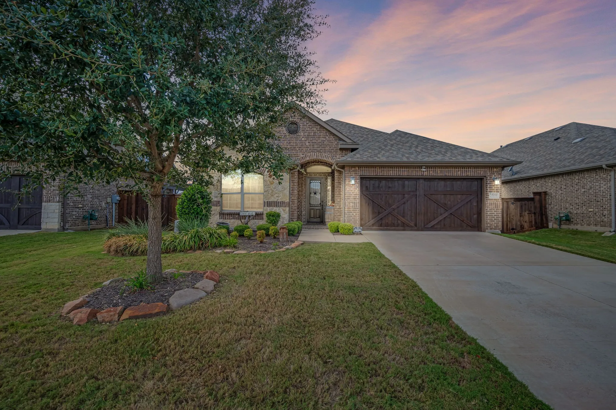 Brick exterior with decorative wood-finish garage doors, stone-bordered landscaping, and paved driveway