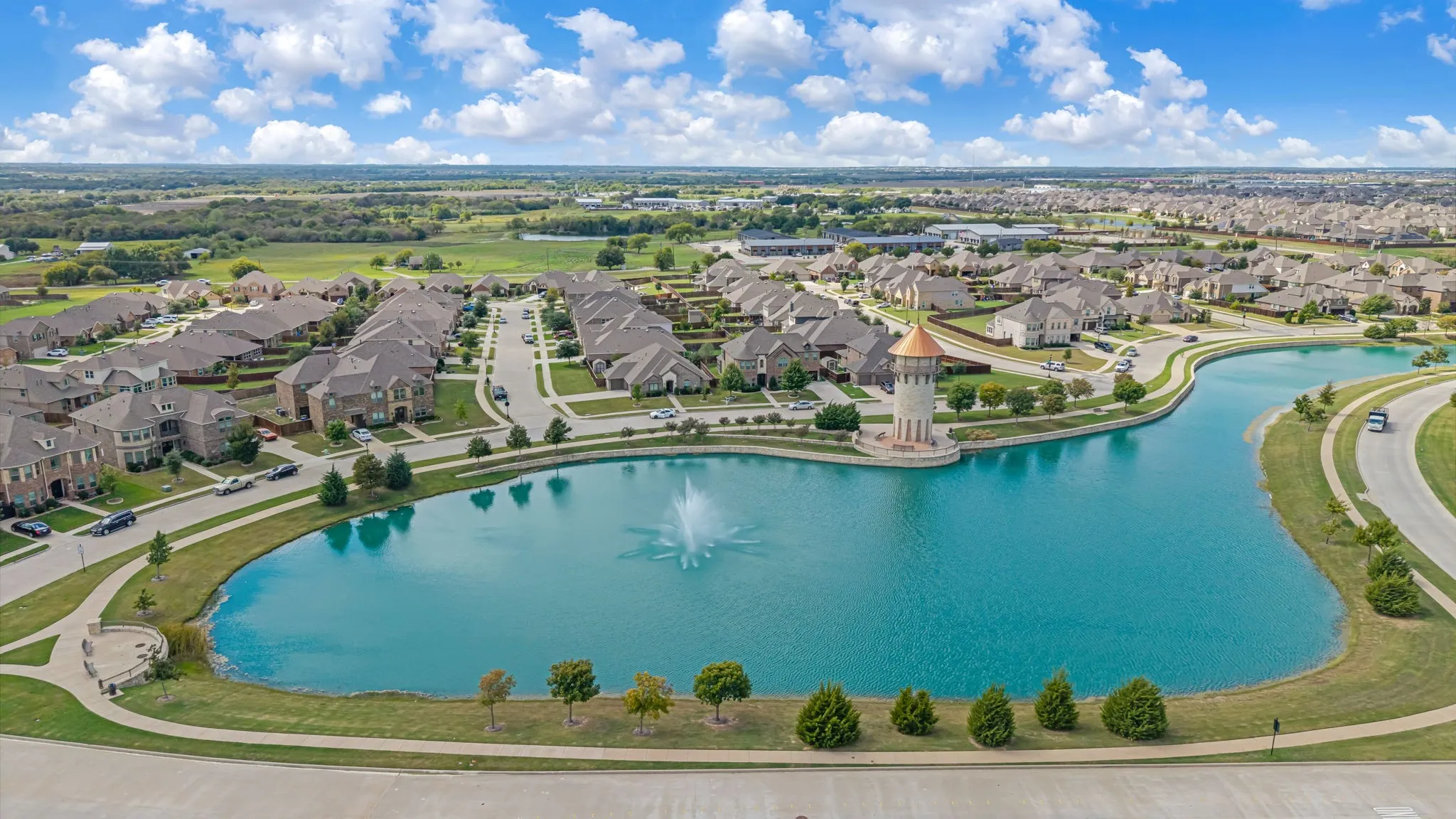 Aerial perspective of the residential community featuring a central pond with a fountain, a stone tower structure, and paved walking paths
