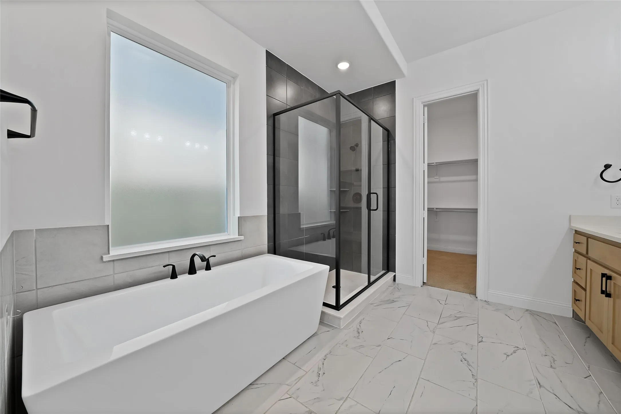 Freestanding soaking tub with matte black fixtures, walk-in shower featuring dark tile surround and glass enclosure, tile flooring, light wood cabinetry, and recessed lighting