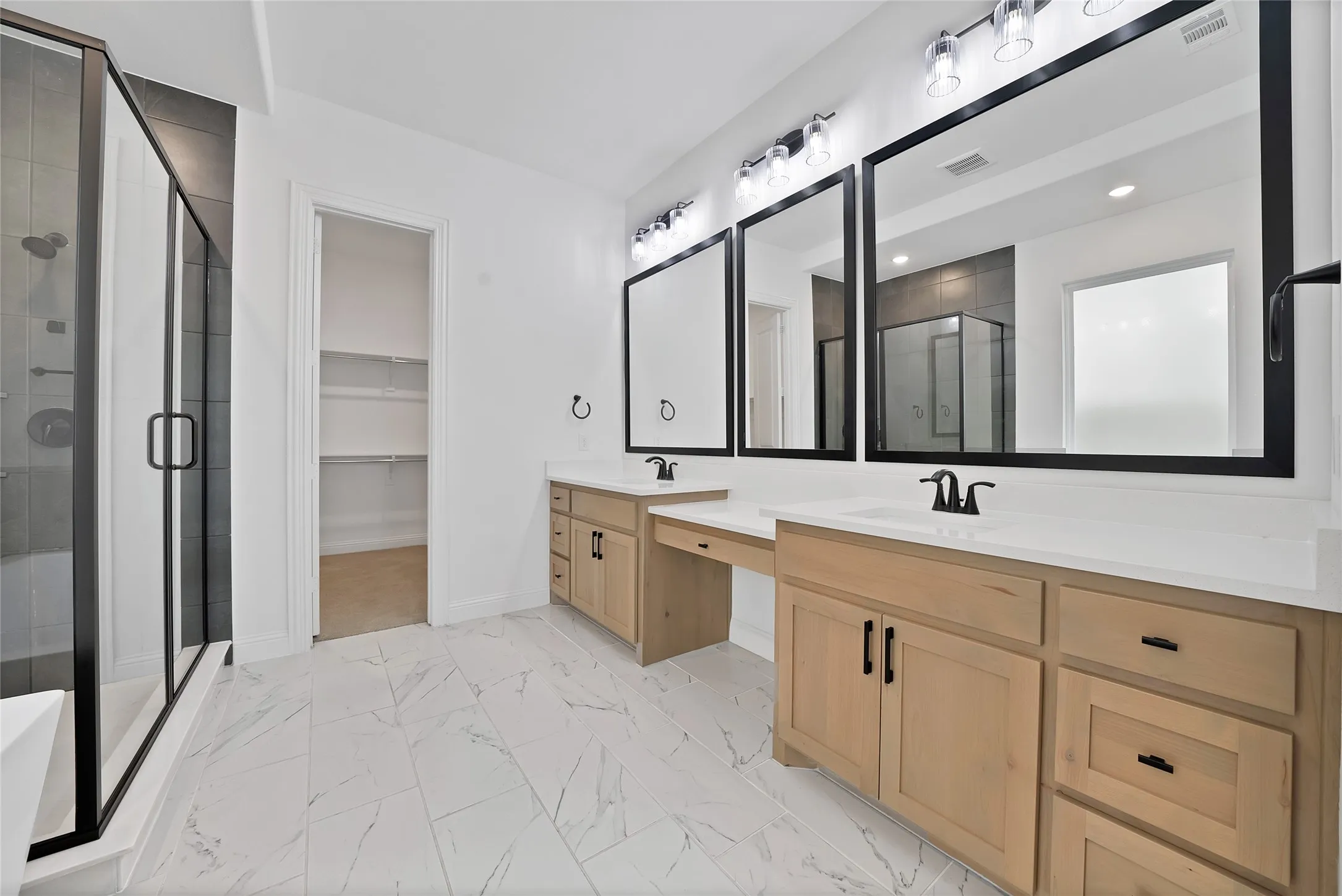 Double vanity featuring light wood-finish cabinetry, white countertops, matte black hardware, and rectangular mirrors, complemented by large-format tile flooring and a glass-enclosed shower