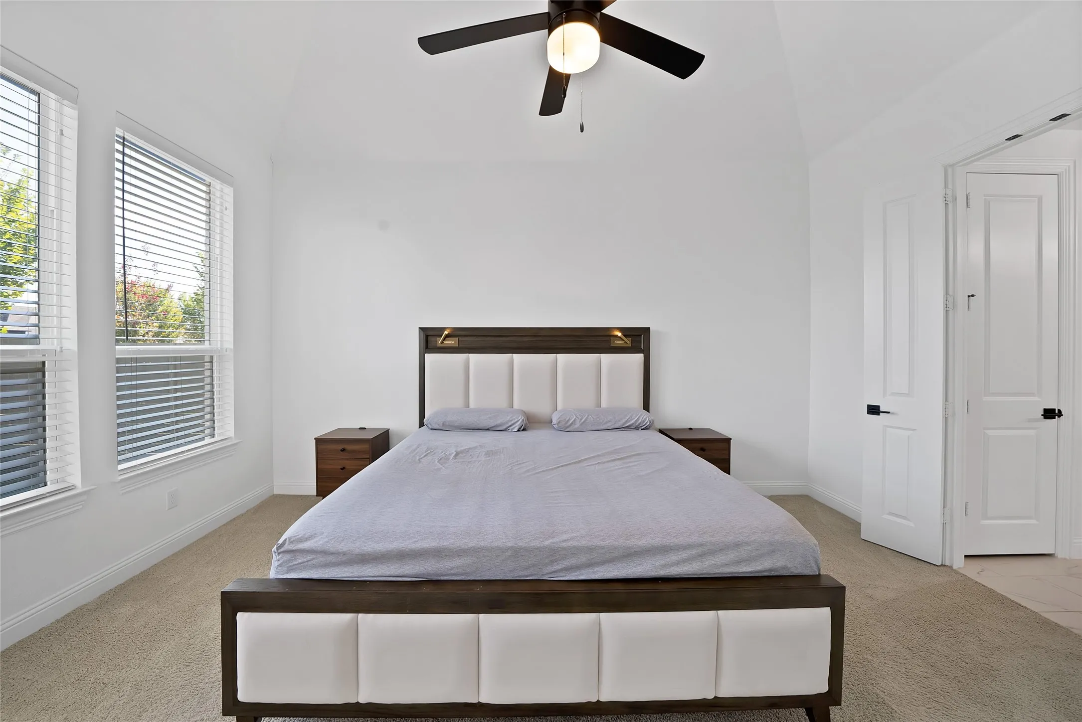 Vaulted ceiling with an integrated matte black ceiling fan, dual windows with horizontal blinds, neutral wall-to-wall carpeting, and paneled doors with matte black hardware