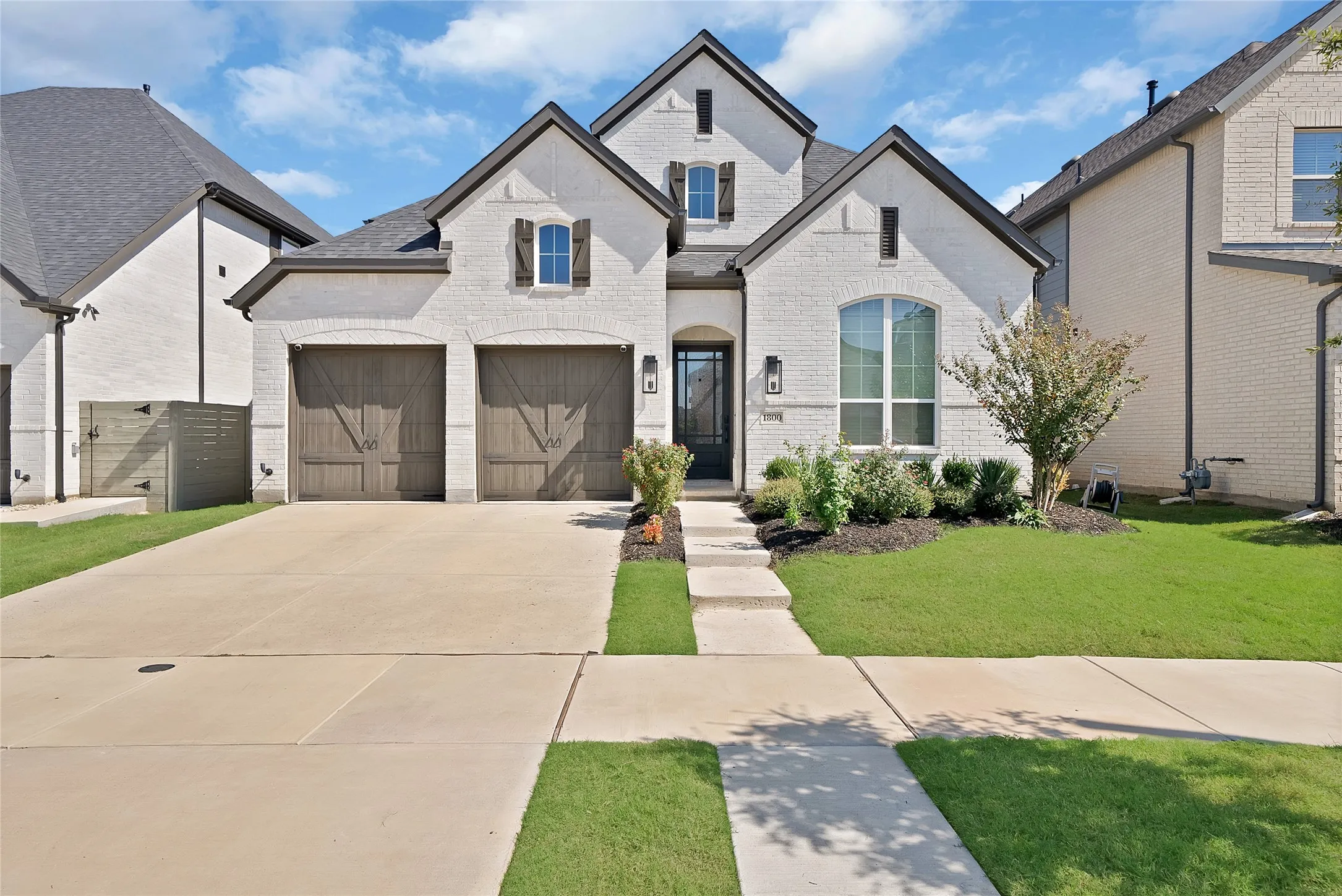 Exterior facade featuring white brick, dark wood-finish carriage-style garage doors, decorative shutters, and a covered entry with a glass-paneled door