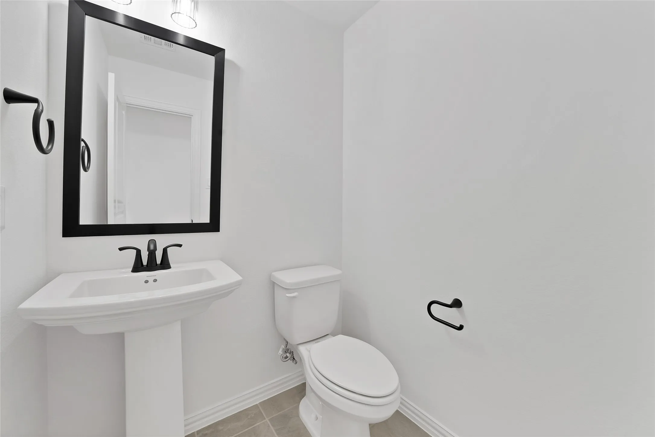 Pedestal sink with a matte black faucet, framed mirror, and tile flooring