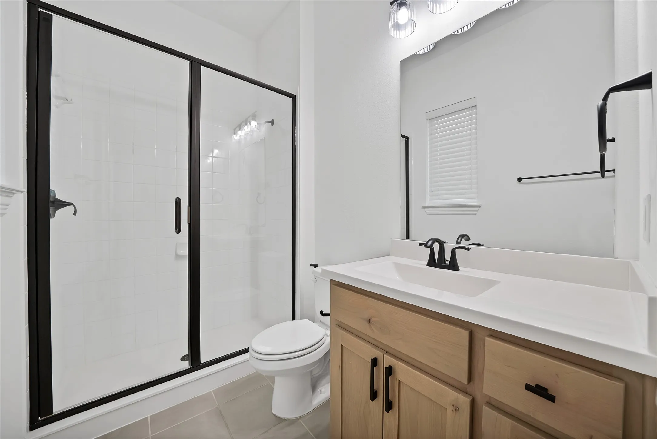 Bathroom featuring a wood-finish vanity with a white countertop, matte black hardware, a glass-enclosed shower, and large-format tile flooring