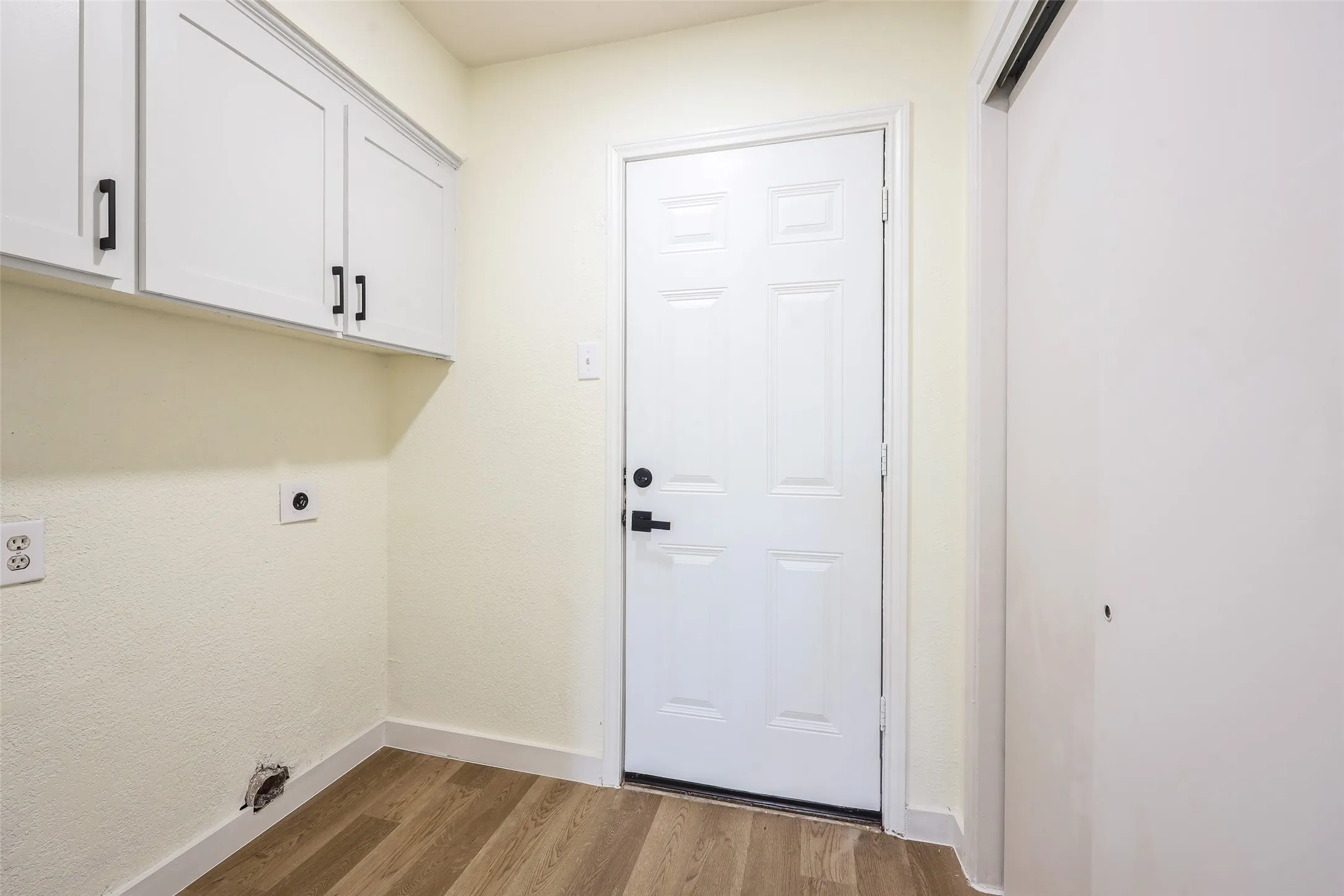 Wood-finish flooring with white baseboards, white paneled door with black hardware, and white upper cabinetry with black pulls
