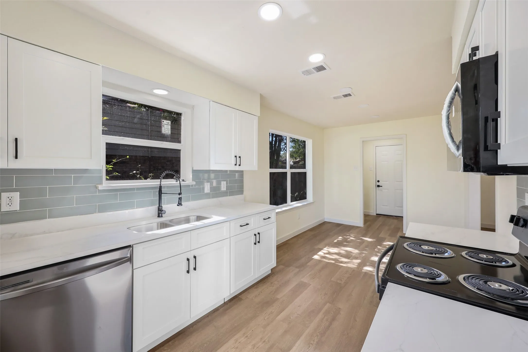 Kitchen featuring white shaker cabinetry, light-toned wood-finish flooring, undermount dual-basin sink with a matte black faucet, stainless steel dishwasher, and subway tile backsplash