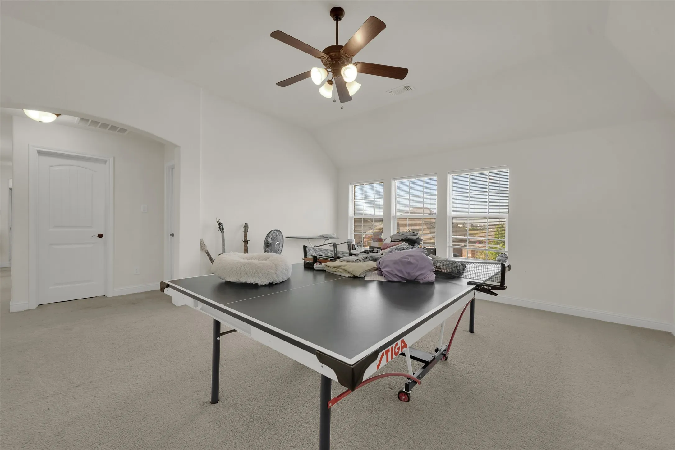 Vaulted ceiling featuring a central multi-blade ceiling fan with integrated light fixture, carpeted flooring, and three-panel grid windows