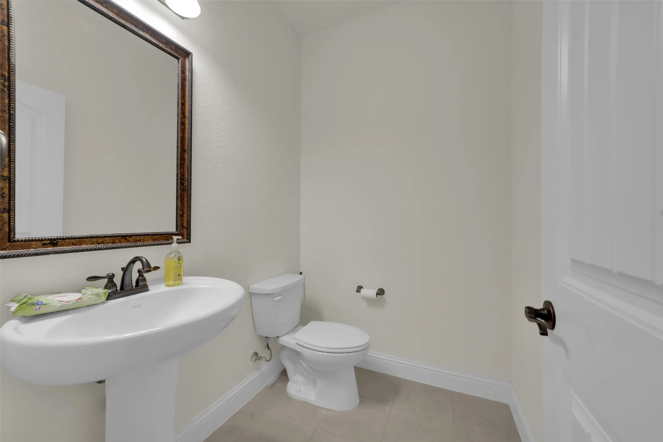 Pedestal sink with dark metal hardware, framed wall-mounted mirror, overhead light fixture, and tile flooring