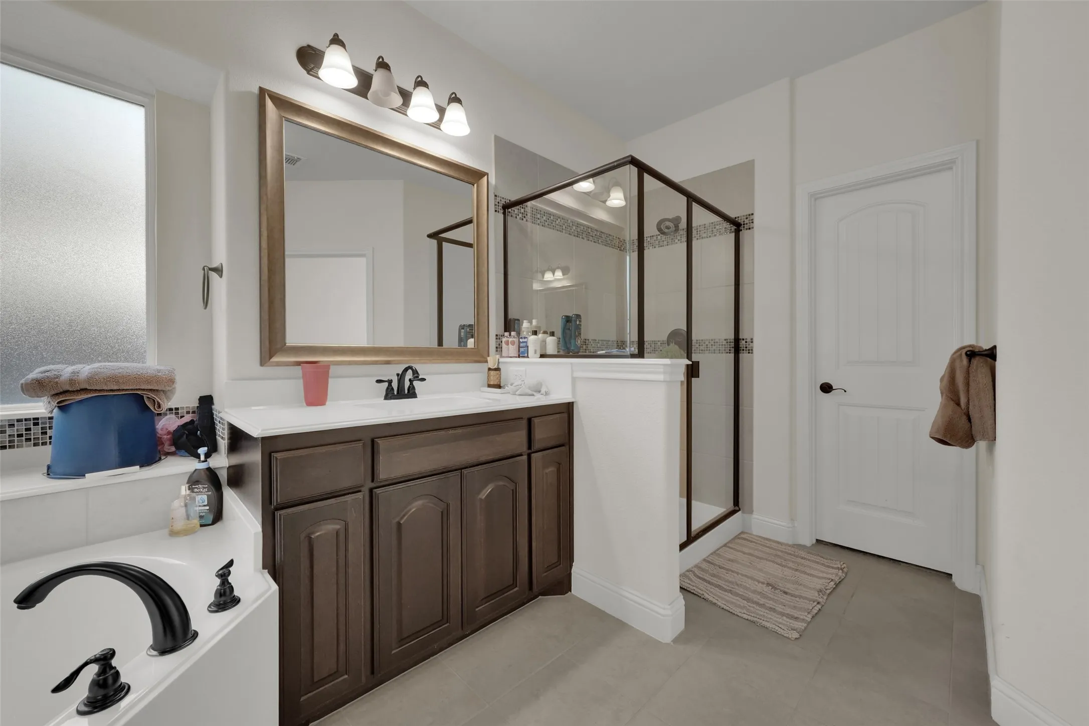 Dark wood vanity with a light-toned countertop, framed wall mirror, four-light vanity fixture, built-in soaking tub, and enclosed glass shower with dark metal framing