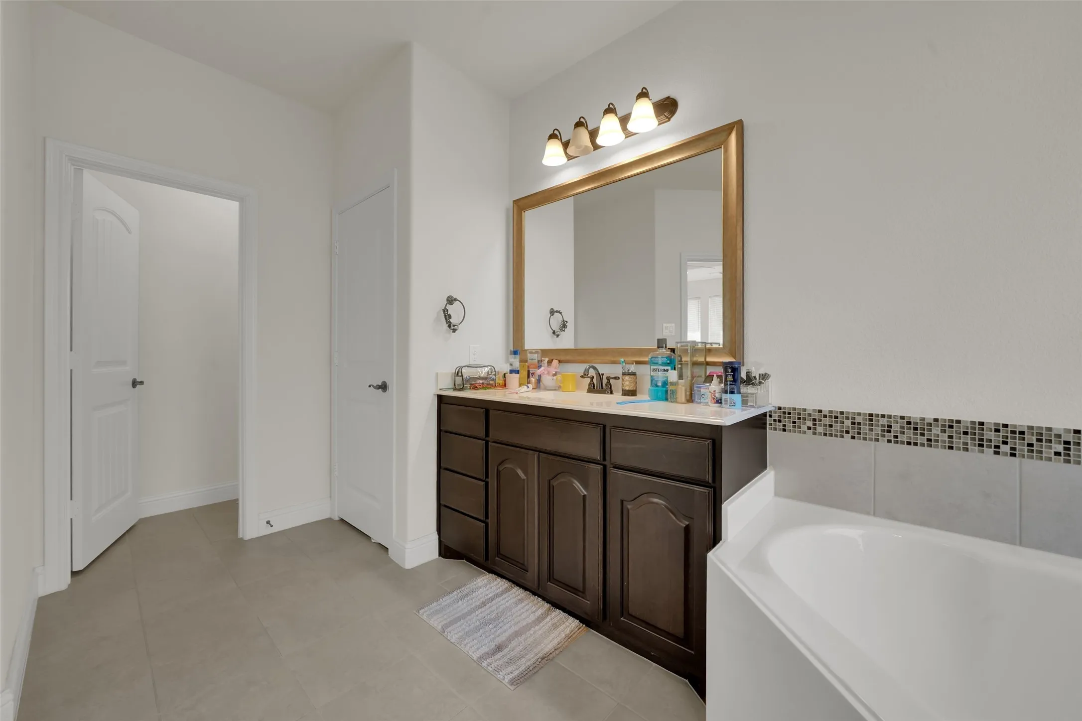 Dark-finish cabinetry vanity with a stone countertop, framed mirror, four-light wall fixture, and soaking tub with decorative tile backsplash