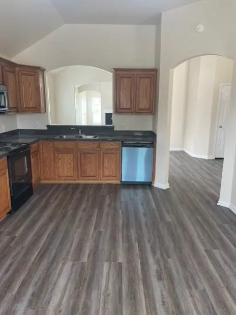 Kitchen featuring wood-finish cabinetry, stainless steel dishwasher, vaulted ceiling, dark solid-surface countertops, and wood-finish flooring