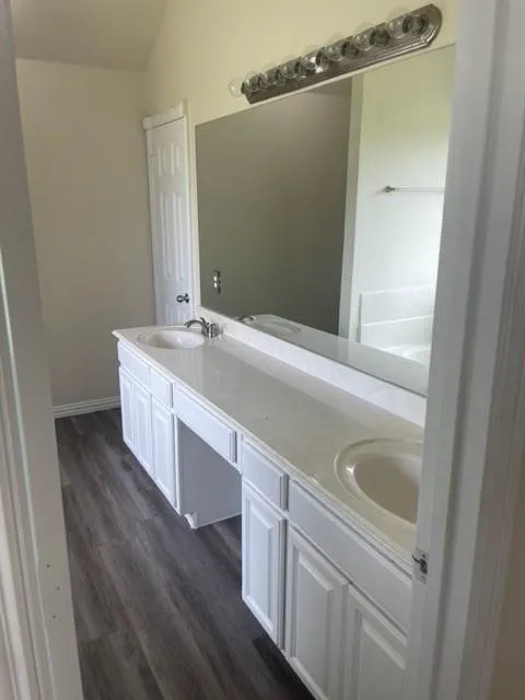 Double vanity featuring white cabinetry, integrated sinks, a large wall-mounted mirror, multi-bulb lighting fixture, and wood-finish flooring