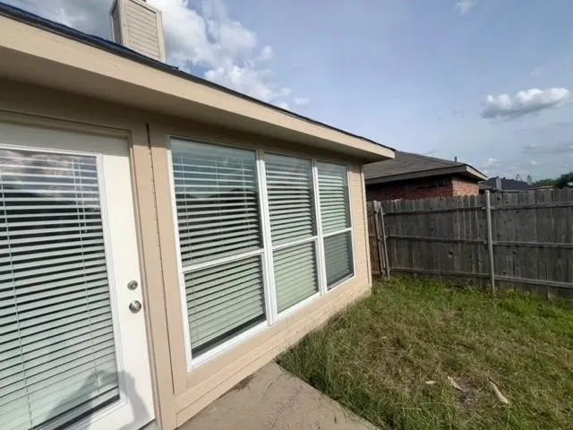 Exterior rear elevation featuring beige siding, white-trimmed windows, a full-lite glass door, concrete hardscaping, and a wood privacy fence