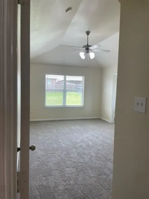 Vaulted ceiling room featuring neutral carpeting, a central multi-blade ceiling fan with integrated lighting, and a window with exterior exposure