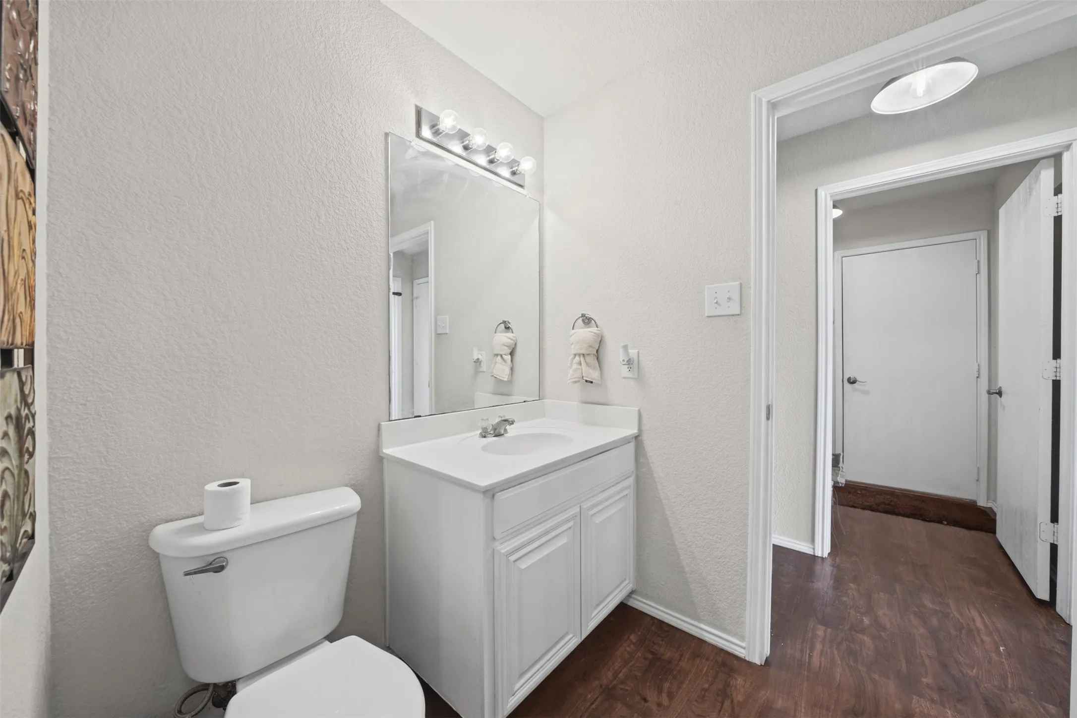 Bathroom featuring a white vanity cabinet with an integrated sink and countertop, large rectangular mirror, four-bulb vanity light, and dark wood-finish flooring