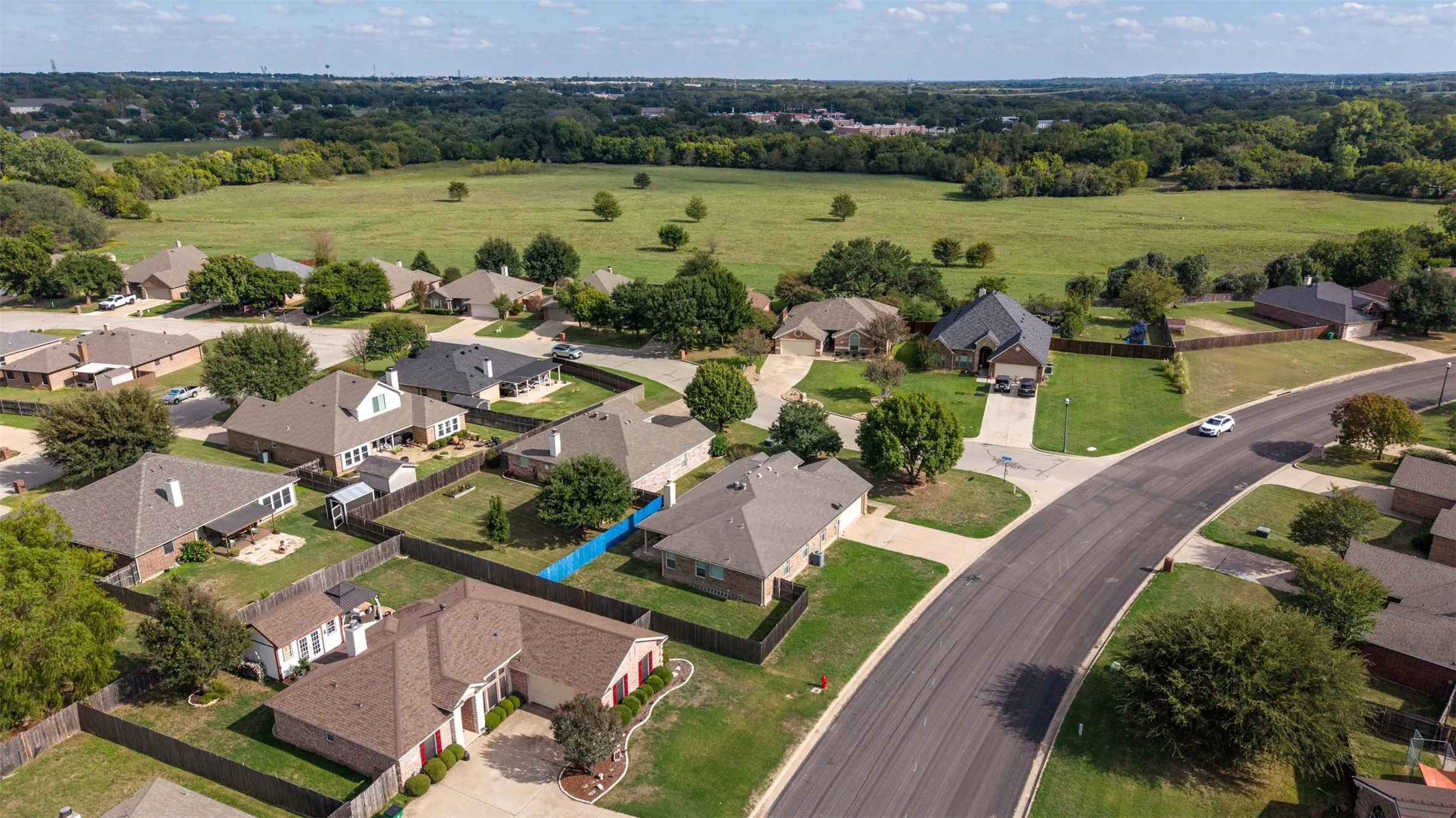 Aerial perspective of residential lots featuring asphalt street access, perimeter wood fencing, mature trees, and expanse of open pastureland