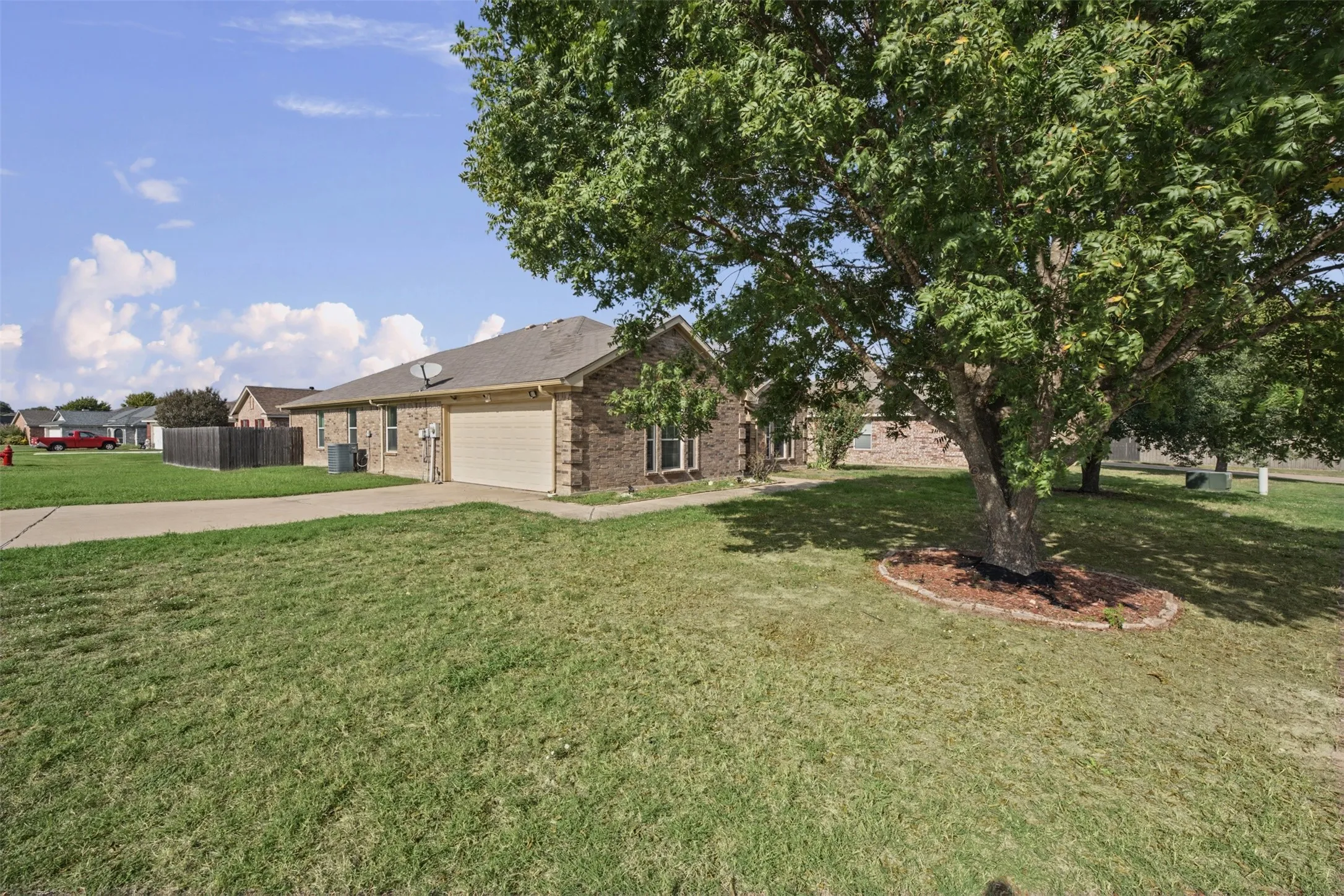 Single-story brick exterior featuring an attached two-car garage, concrete driveway, and expansive front lawn with a mature tree