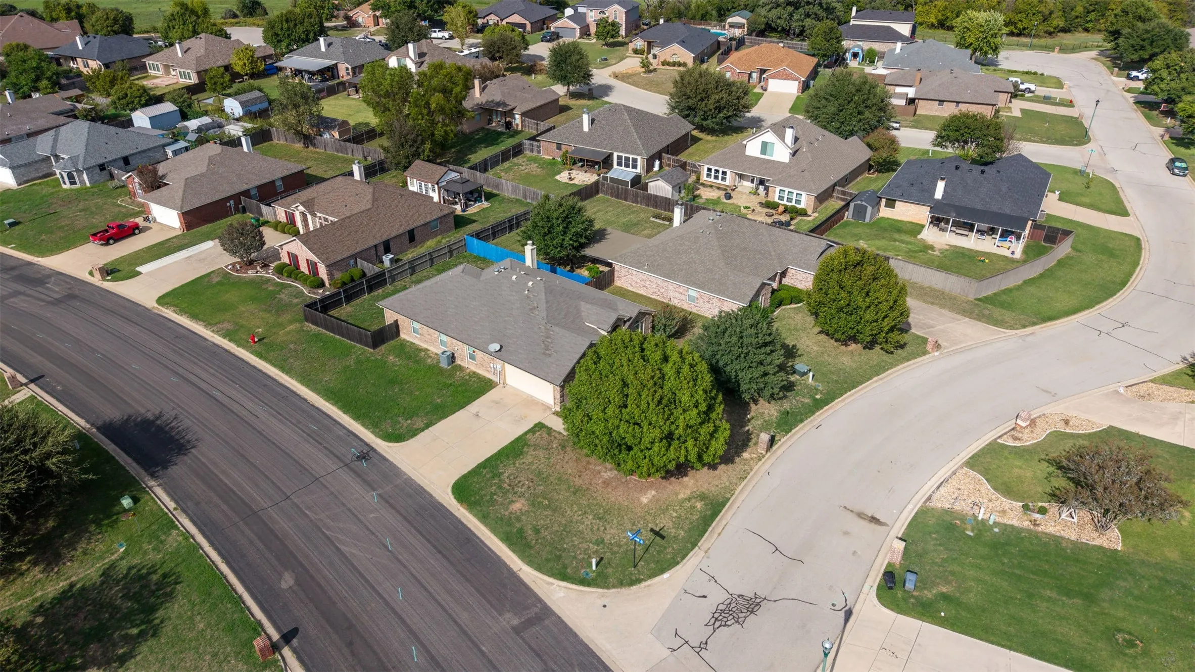 Aerial perspective of a single-story residence featuring a brick and stone facade, a multi-gabled shingle roof, a private fenced backyard, and a concrete driveway