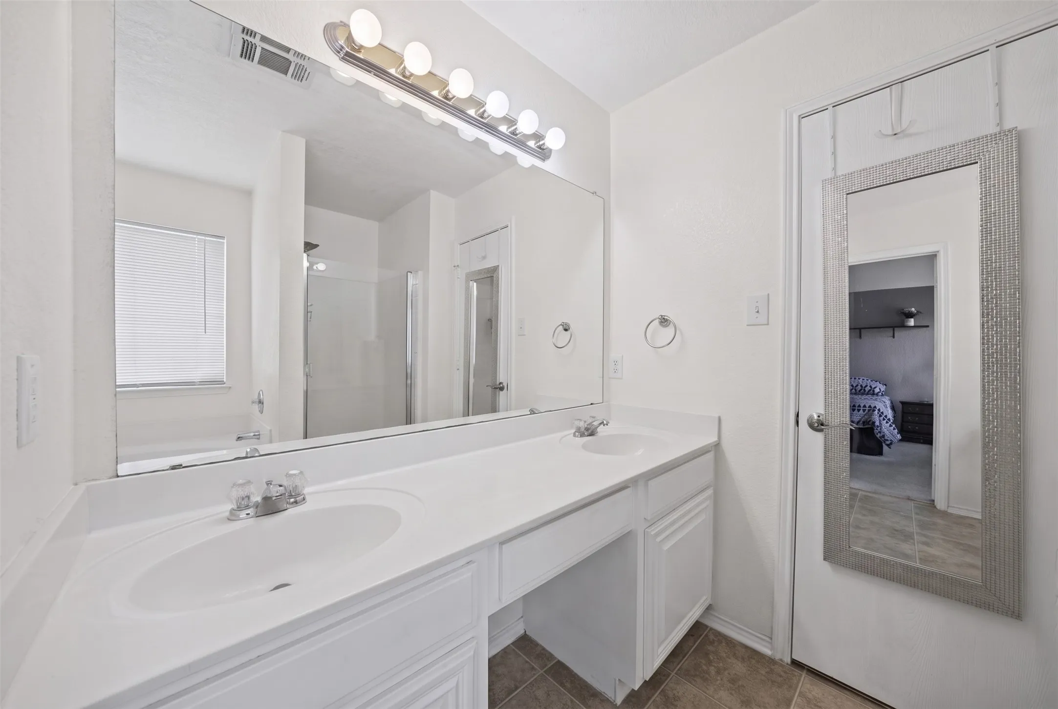 Dual-sink vanity with white cabinetry and solid surface countertop, featuring a large wall-to-wall mirror, multi-bulb overhead lighting, and tile flooring