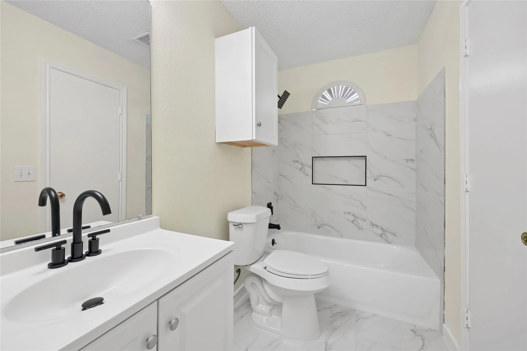Bathroom featuring a white vanity, matte black widespread faucet, stone-patterned tile tub surround with an integrated niche, and an arched transom window