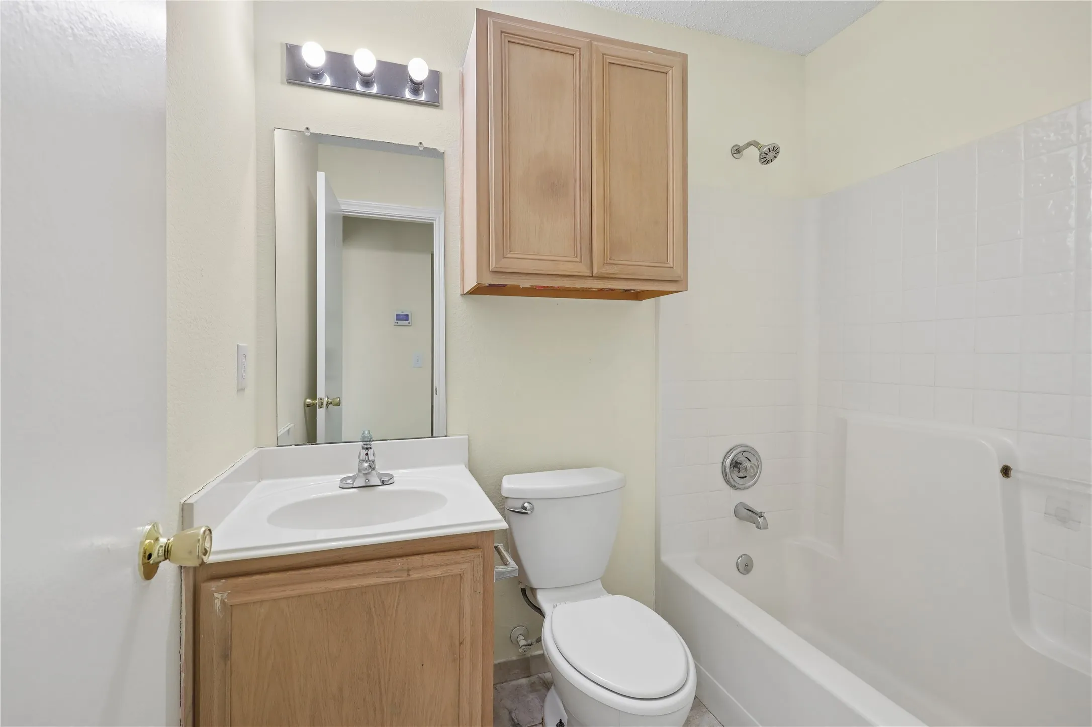 Bathroom featuring a vanity with a white countertop and integrated sink, wall-mounted upper cabinetry, a chrome three-light fixture, a rectangular mirror, and a tub-shower combination with white wall surround
