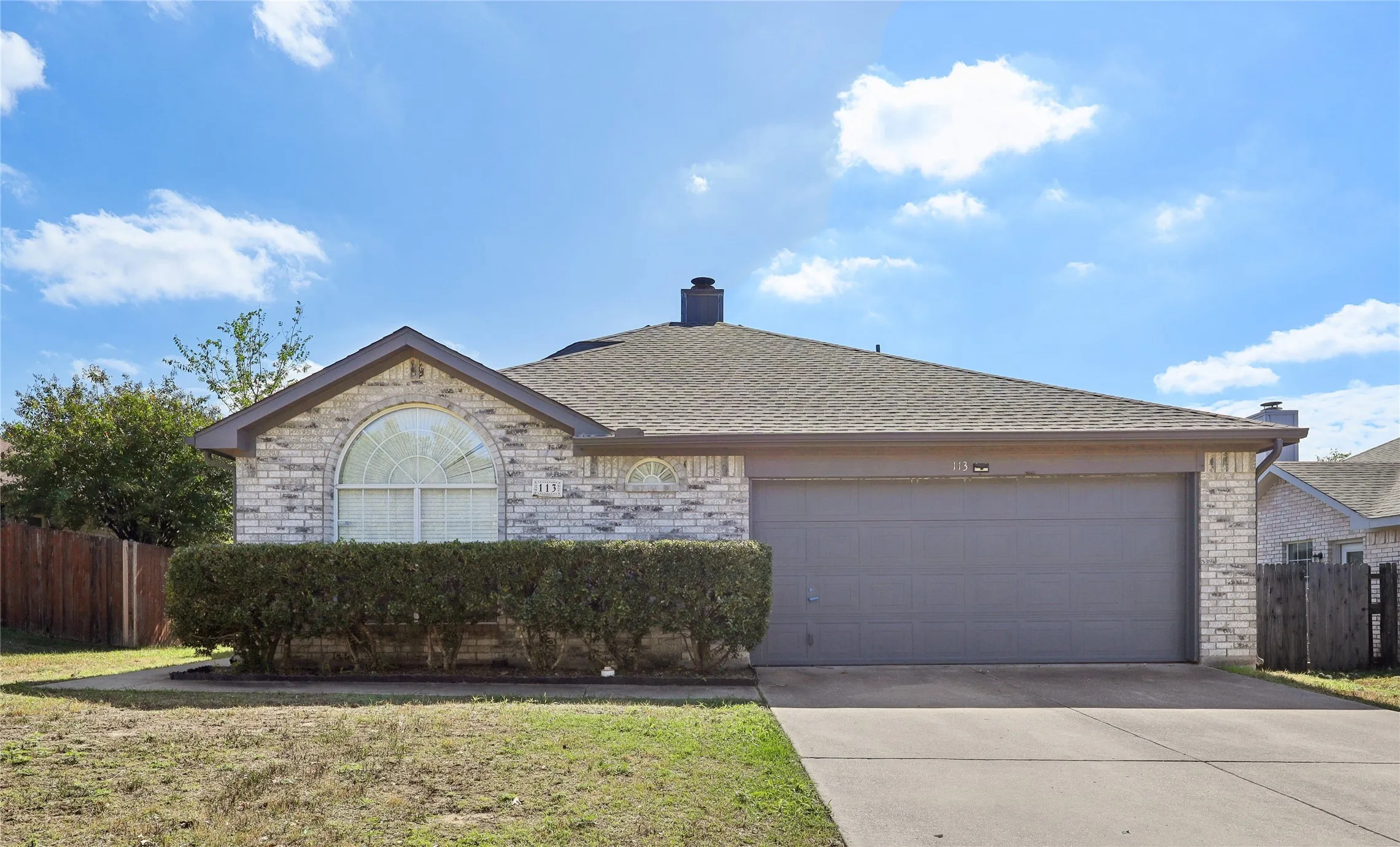 Single-story brick exterior featuring a prominent arched window, shingle roof, two-car garage, and concrete driveway