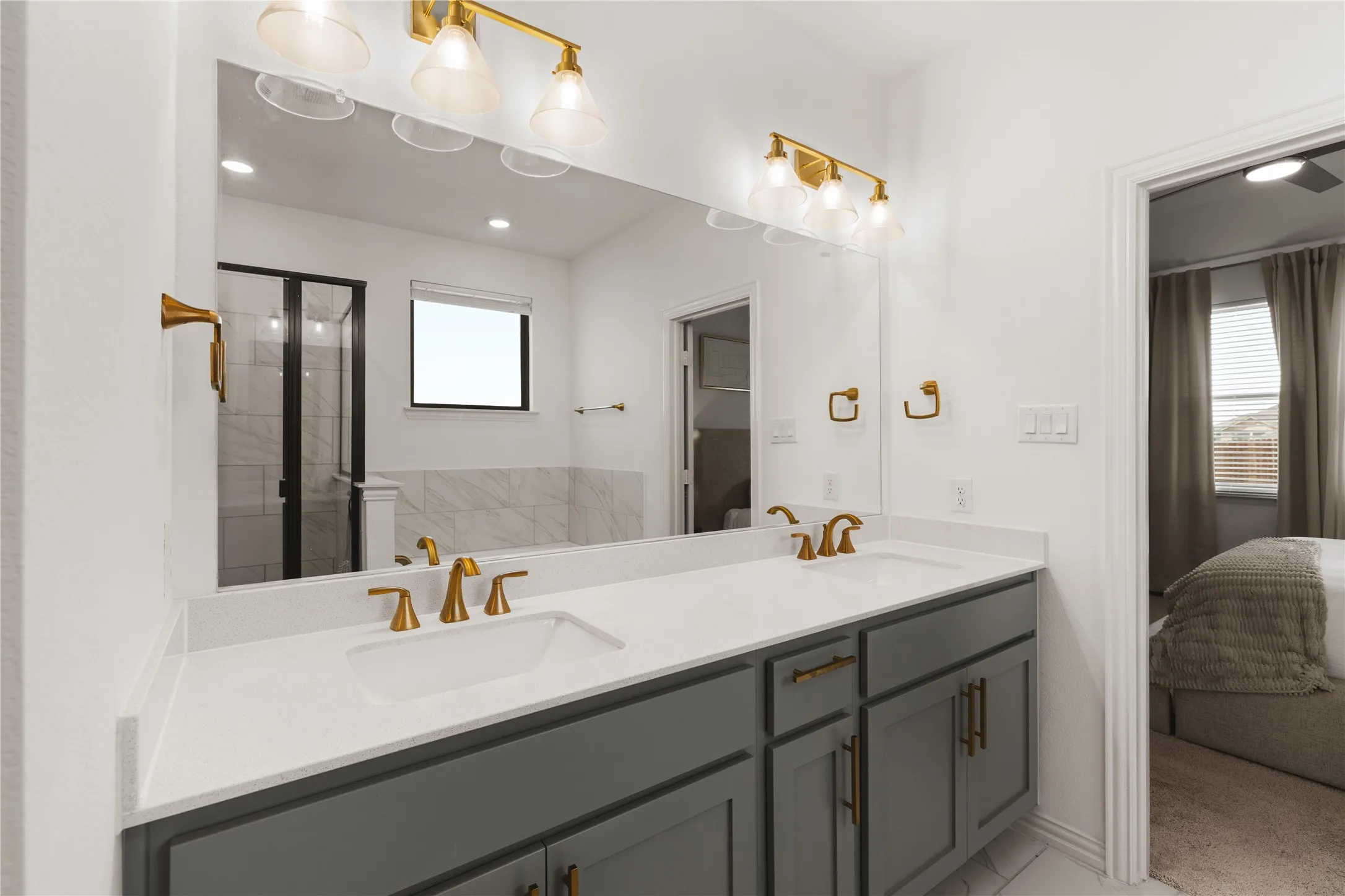 Dual vanity with light-toned solid surface countertops, undermount sinks, brass-finish fixtures, dark cabinetry, and full-width wall mirror
