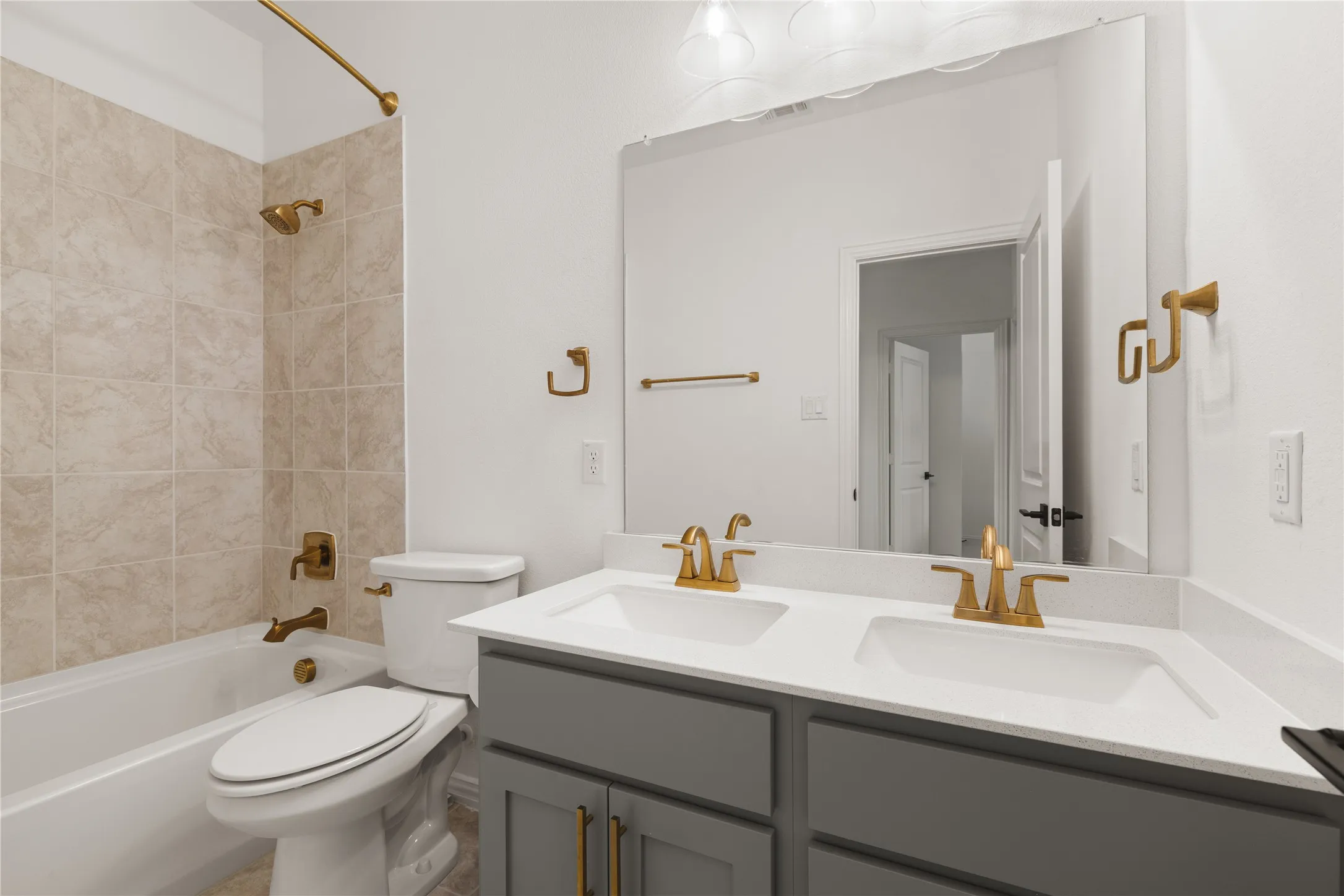 Dual-sink vanity with quartz countertop, grey cabinetry, wall-to-wall mirror, and brass-finish hardware and fixtures, including a tiled shower-tub combination