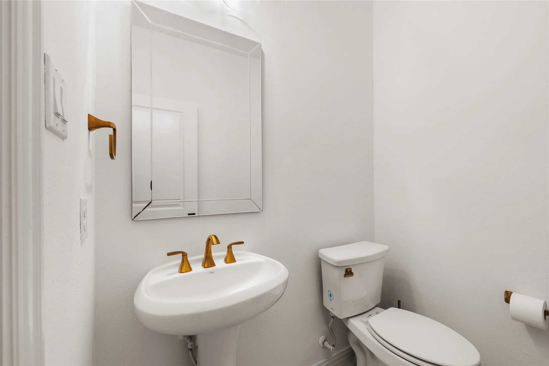 Wall-mounted beveled mirror, white pedestal sink with brass-toned widespread faucet, and matching brass-toned hardware