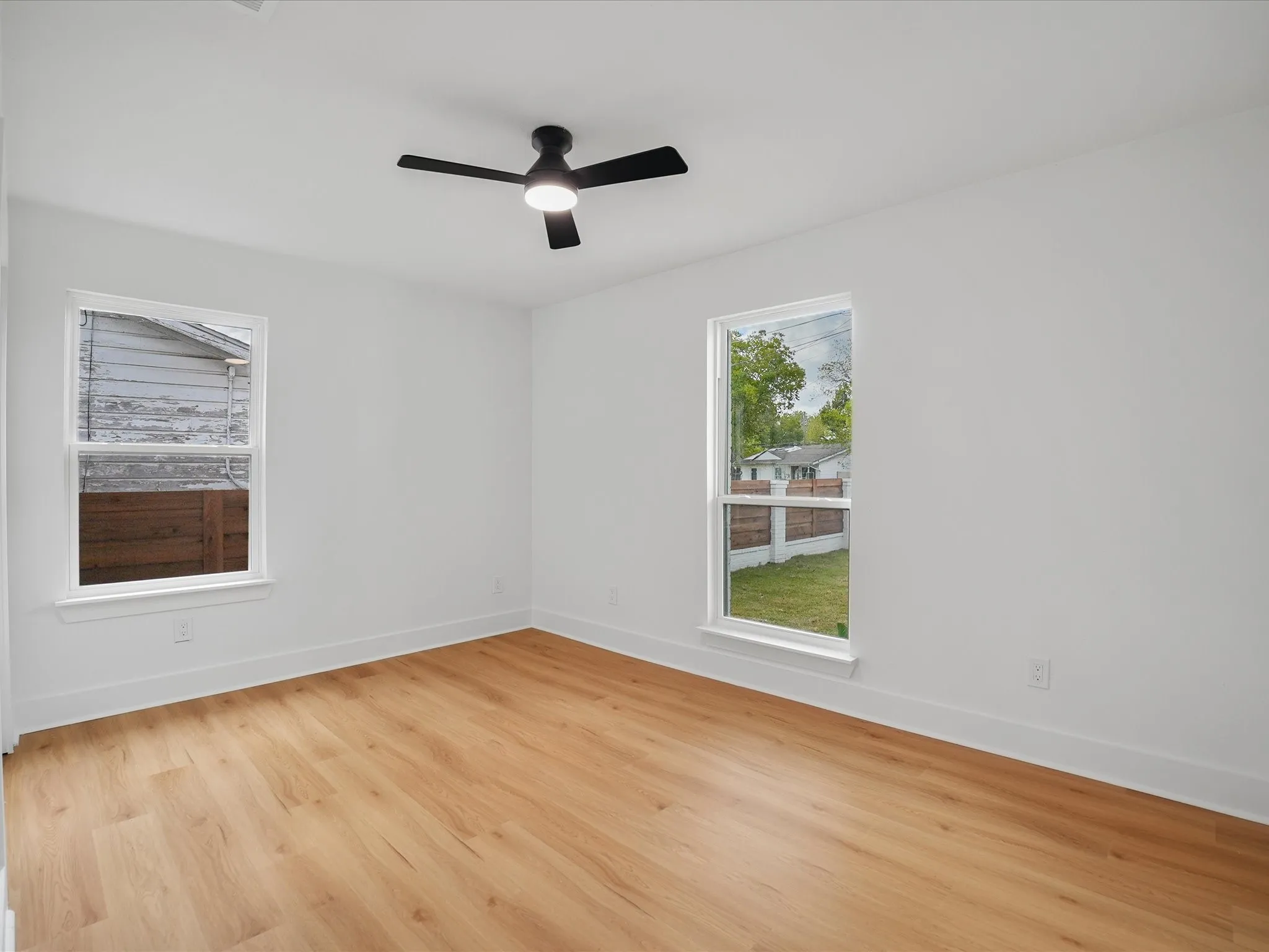 Room featuring wood-finish flooring, white walls, dual windows, and a matte black ceiling fan with integrated lighting