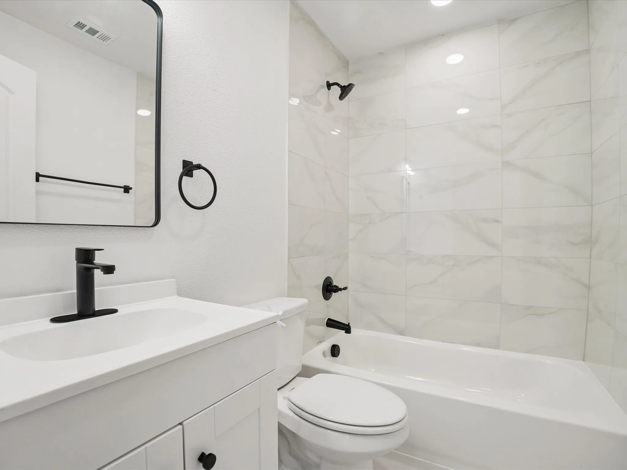 Bathroom featuring a white vanity with a solid surface countertop, matte black faucet, rectangular mirror with a black frame, and a tub-shower combination with floor-to-ceiling neutral-toned tile surround