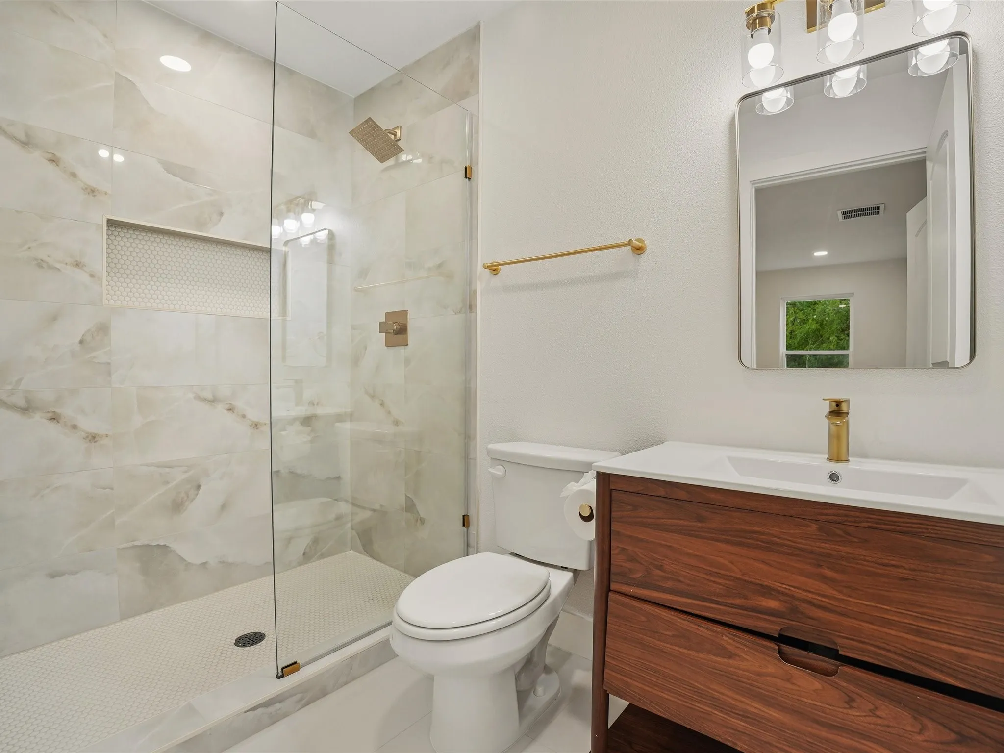 Bathroom featuring a wood-finish vanity with a white countertop, walk-in shower with large-format tile walls and a built-in recessed niche, gold-toned hardware, and a modern rectangular mirror