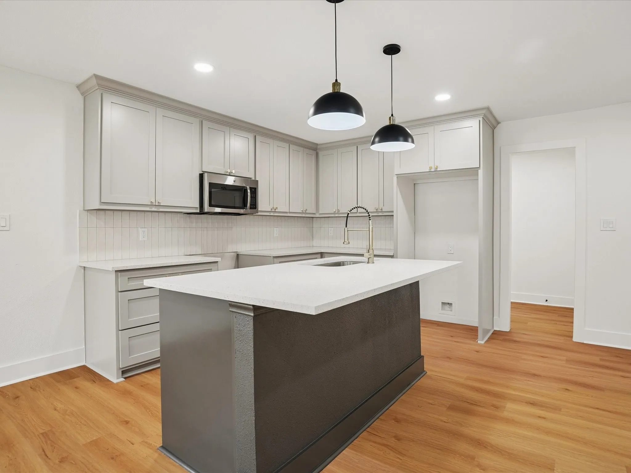 Kitchen featuring light gray cabinetry, solid-surface countertops, a center island with a built-in sink, wood-finish flooring, and two dome-style pendant lights