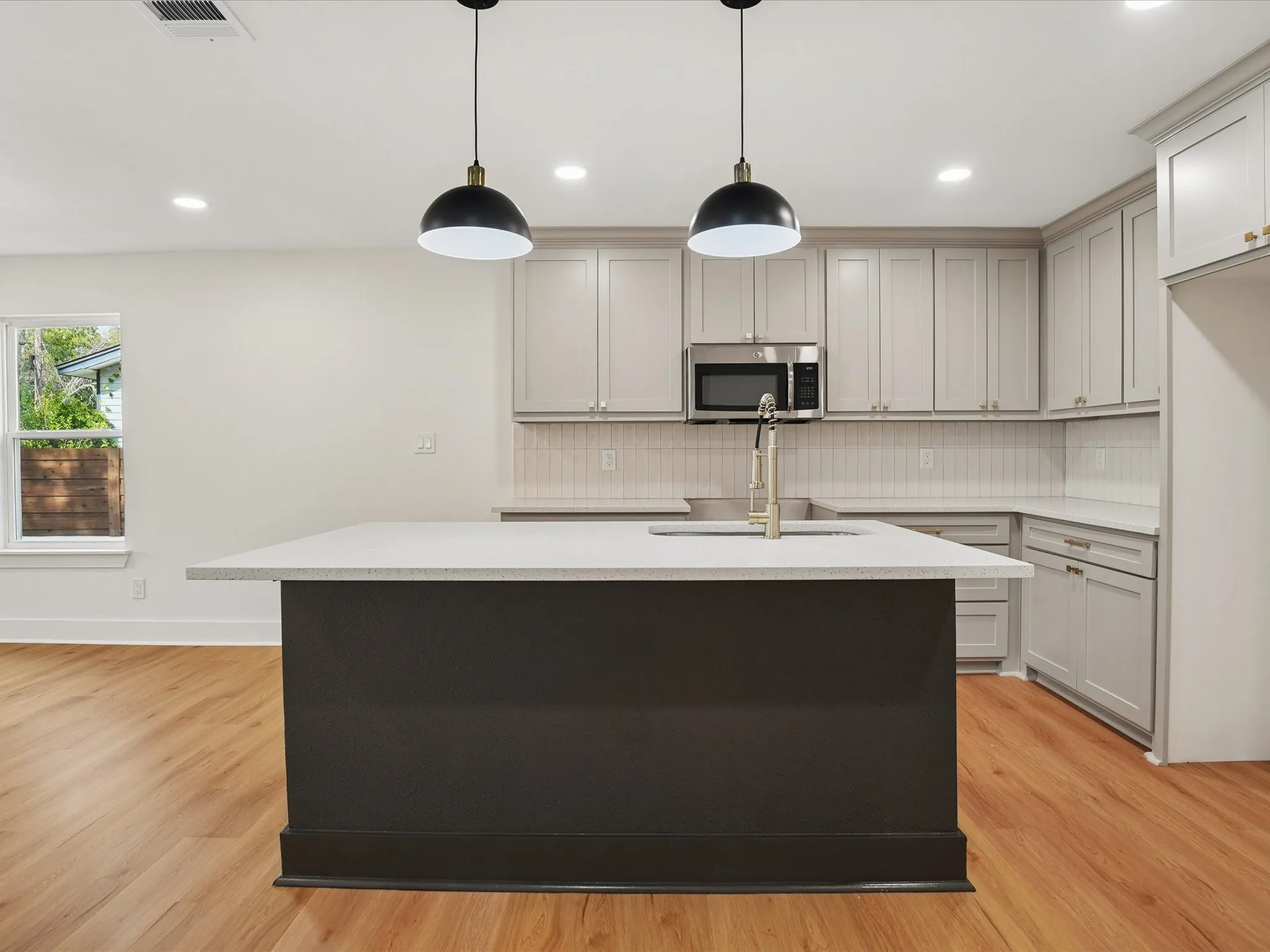Kitchen featuring a central island with a solid surface countertop, light-toned cabinetry, stainless steel microwave, vertical-tile backsplash, and dual pendant lighting over wood-finish flooring