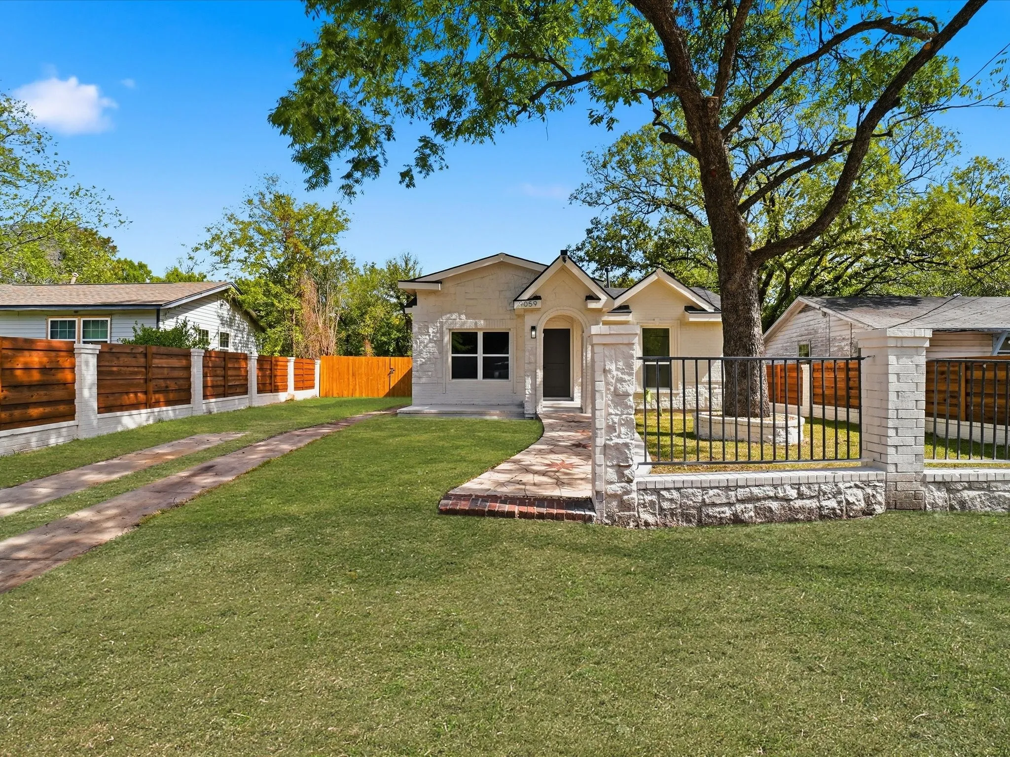 Light-colored masonry exterior with a gabled roofline, stone perimeter fencing, wrought iron gated entrance, and a paved walkway leading to the front entry