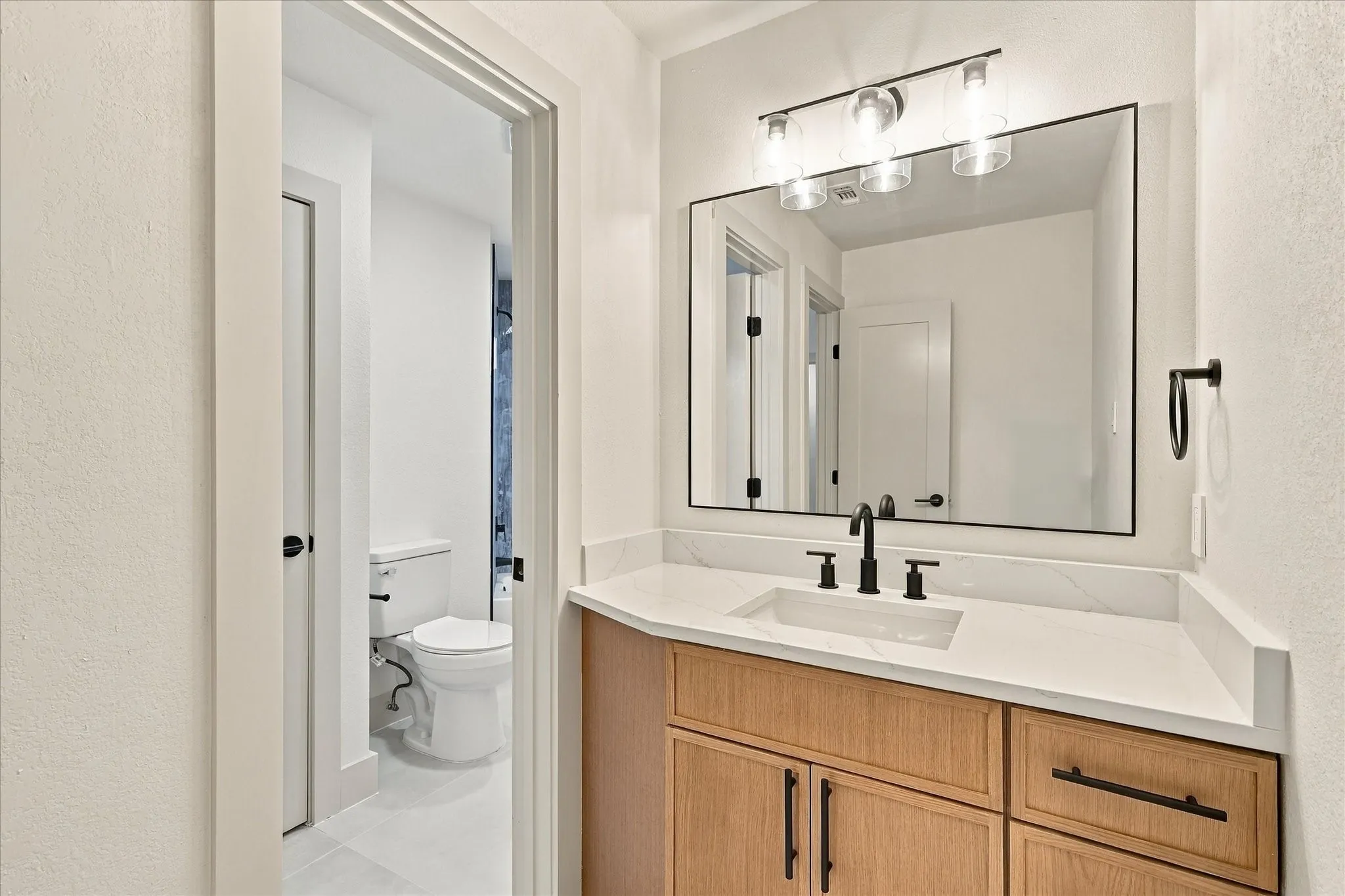 Bathroom vanity featuring a light wood-finish cabinet, white countertop with integrated rectangular sink, matte black widespread faucet, and a modern three-light fixture above a rectangular mirror