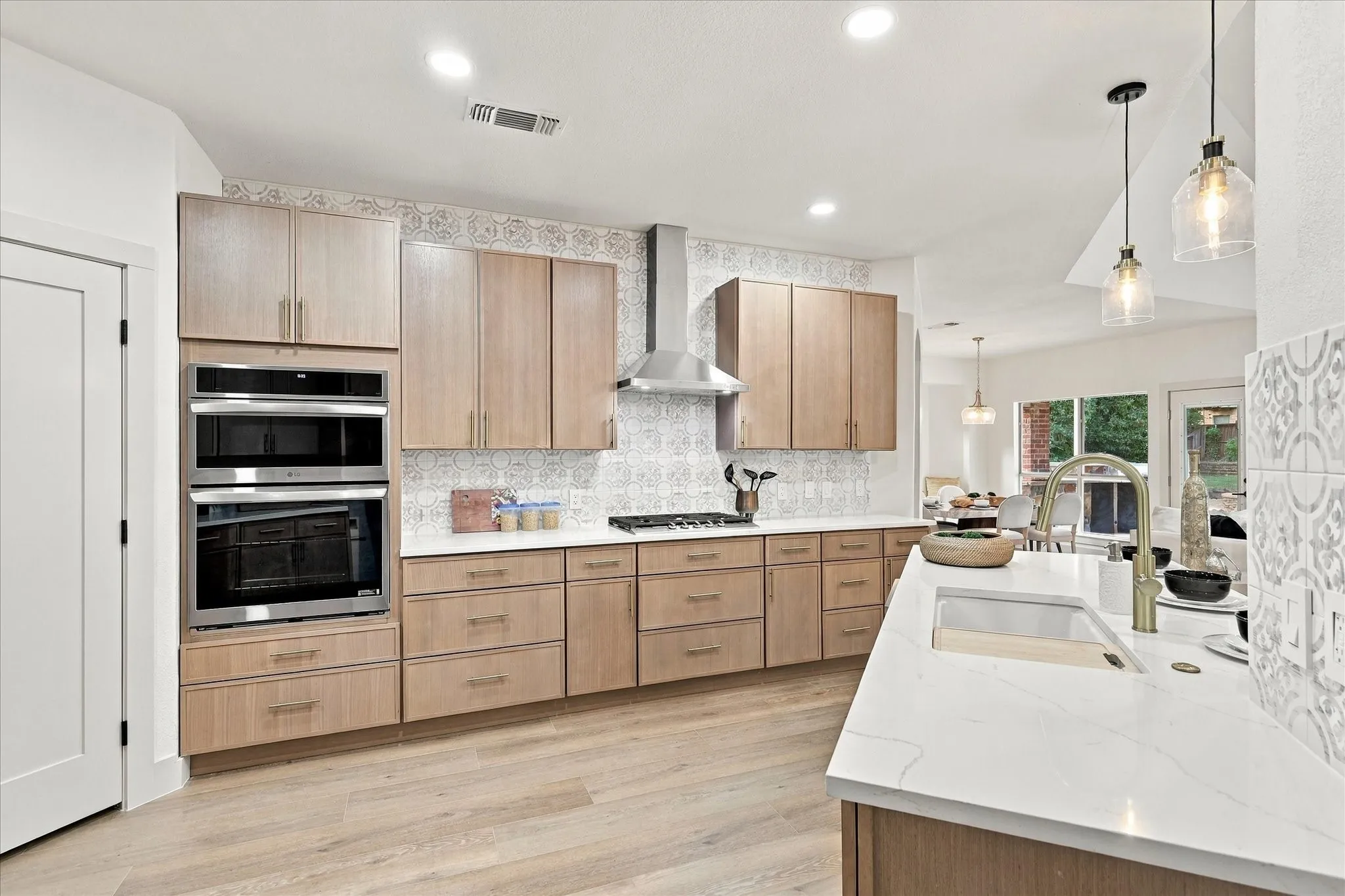 Kitchen featuring light-finish cabinetry, white countertops with veining, patterned tile backsplash, stainless steel built-in double oven and range hood, and wood-finish flooring