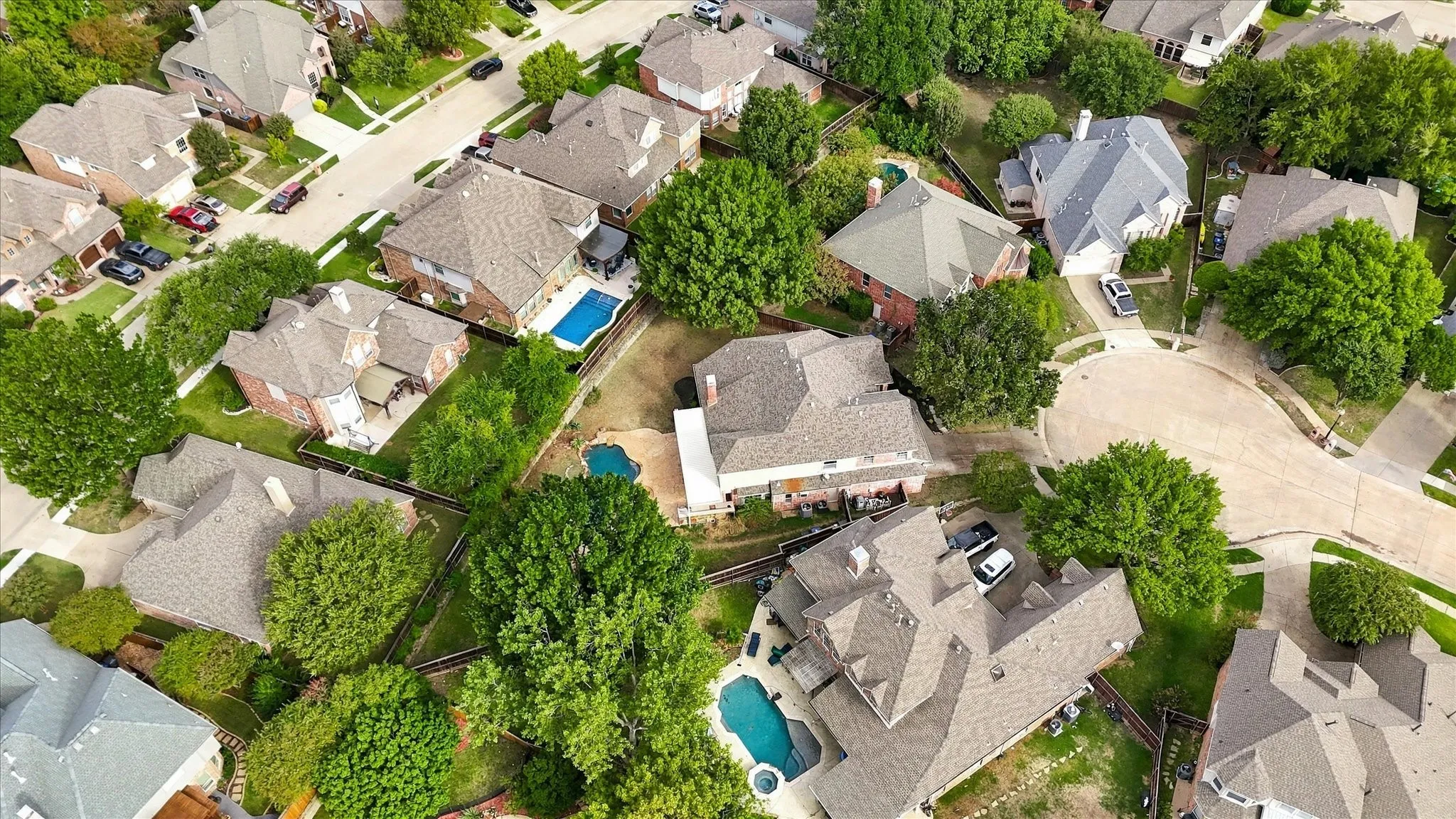 Aerial perspective of a multi-story residence featuring a shingled roof, a backyard in-ground swimming pool, an attached garage, and mature tree landscaping