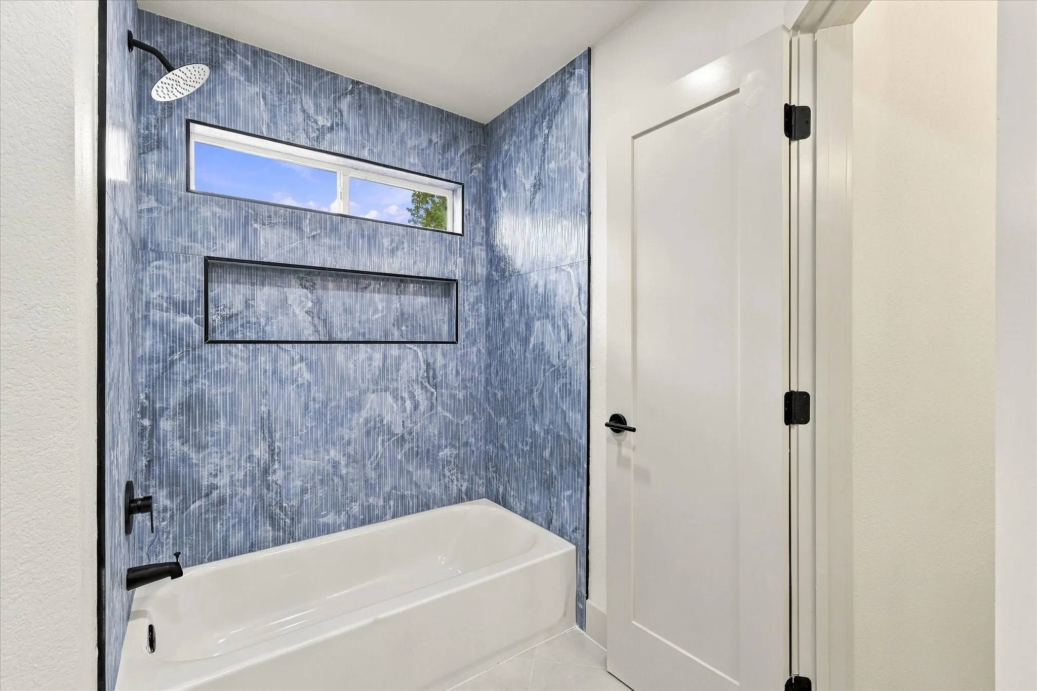 Bathtub surround featuring blue patterned tile, matte black fixtures, an integrated wall niche, and a transom window