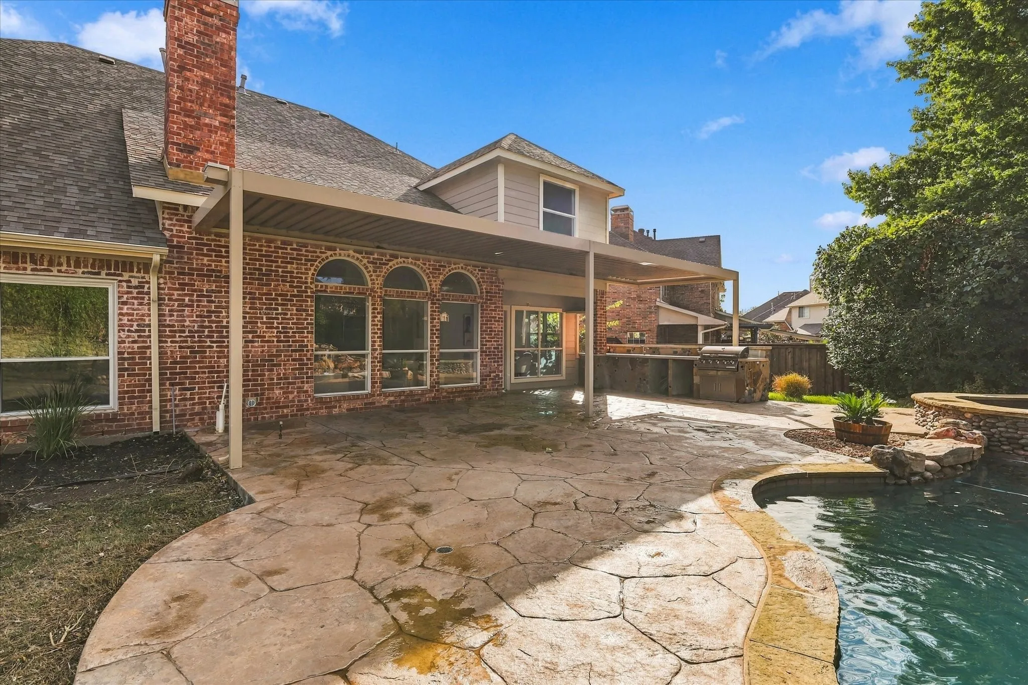 Brick exterior with arched windows, covered patio, outdoor kitchen with stone finish, and swimming pool