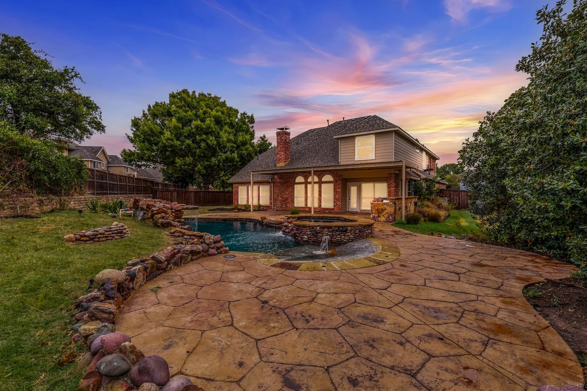 Rear exterior featuring a brick and siding facade, covered patio, stone-accented pool with integrated spa, and extensive flagstone hardscaping