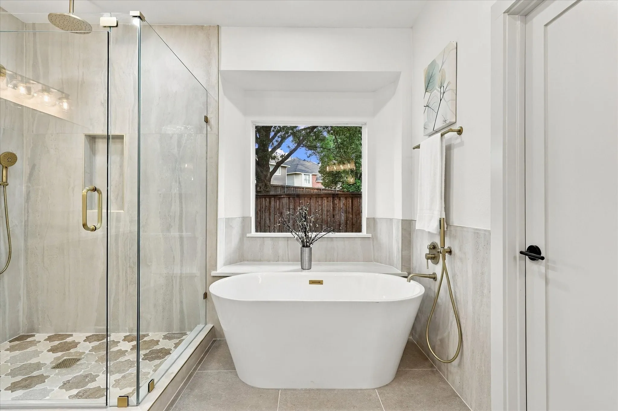 Freestanding soaking tub with brass fixtures, frameless glass shower enclosure featuring an integrated stone-tiled niche, patterned tile shower flooring, and oversized window with stone-topped ledge