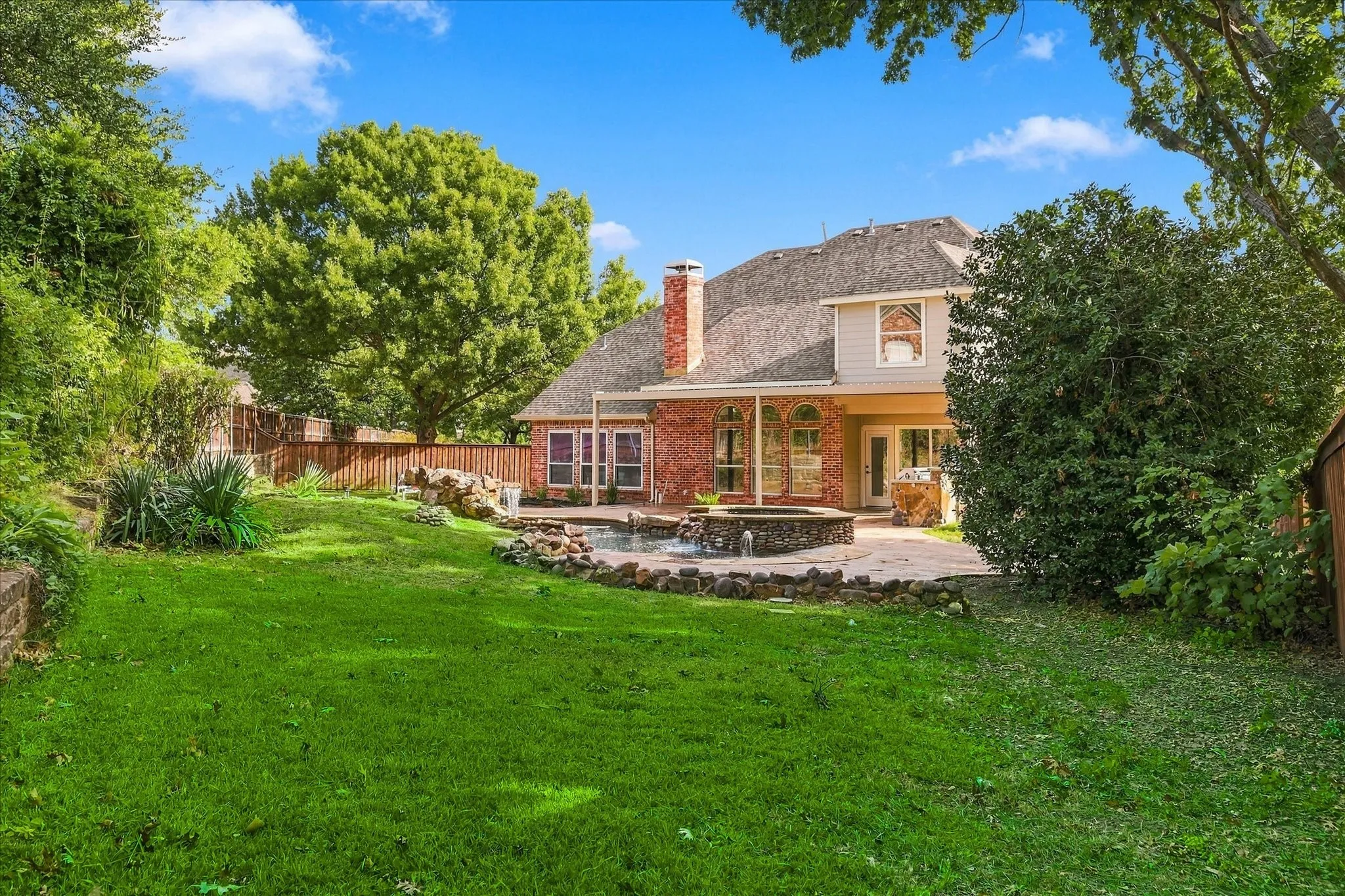 Backyard featuring a brick and siding exterior, stone-accented spa, concrete patio, and mature trees enclosed by a wooden privacy fence