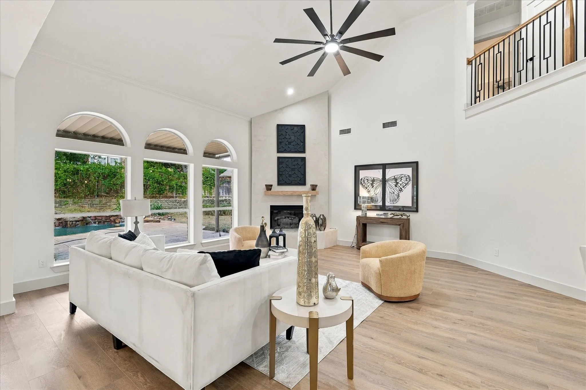 Vaulted ceilings with a multi-blade ceiling fan, expansive arched windows, wood-finish flooring, and a fireplace with a stone-surround facade featuring a wooden mantel