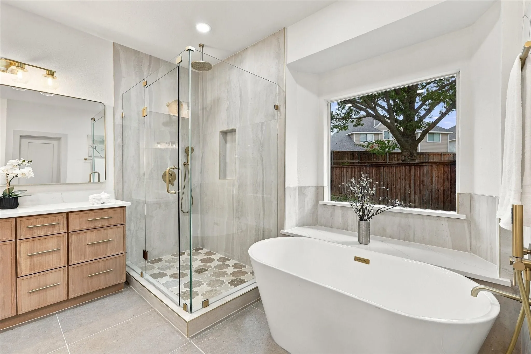 Freestanding soaking tub, glass-enclosed walk-in shower with stone tile surround, patterned tile shower floor, light wood-finish vanity, and picture window