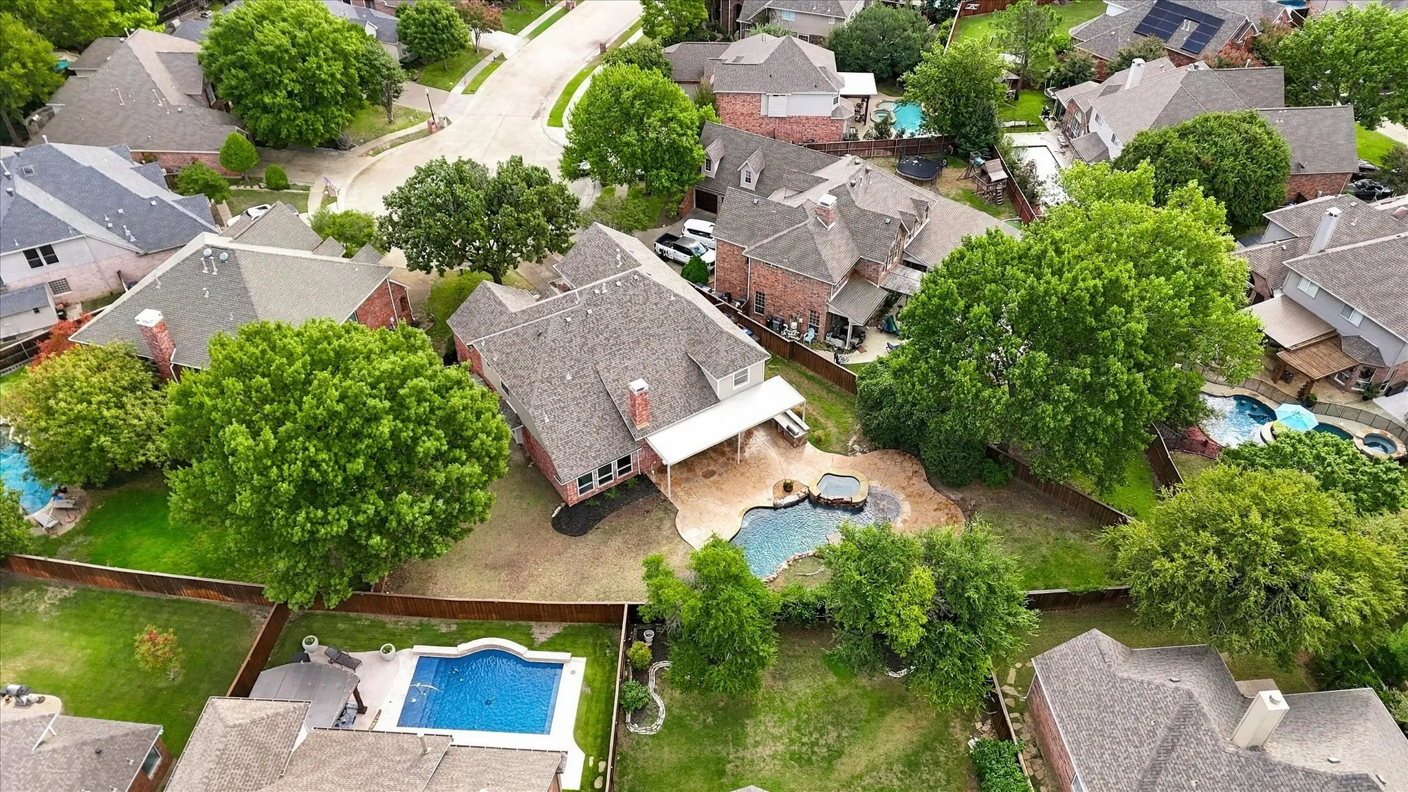 Aerial view of a residential property featuring a brick exterior, a covered patio, an in-ground swimming pool, a separate spa, and mature trees