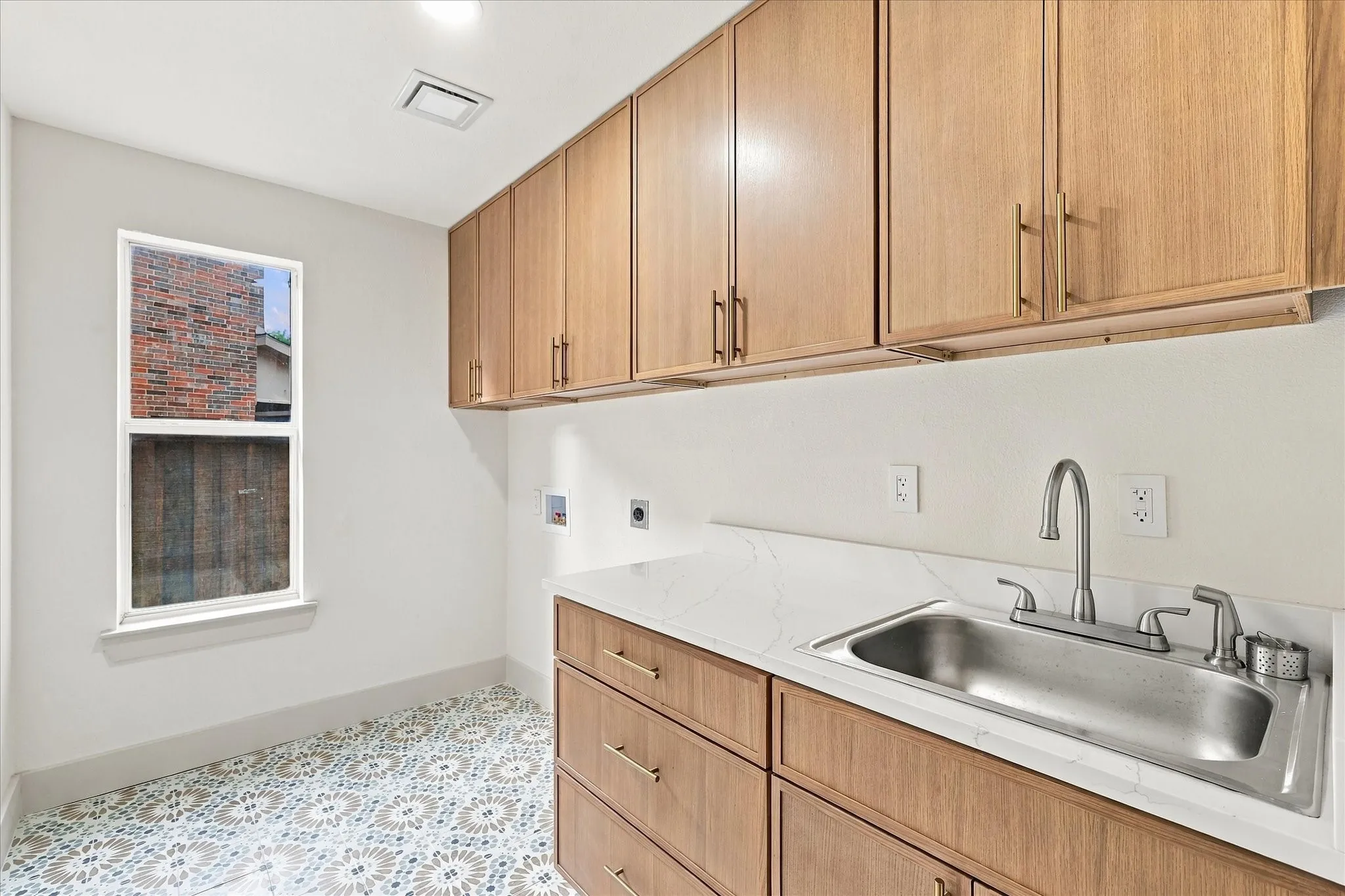 Laundry area featuring natural wood-finish cabinetry, stone-surface countertop, stainless steel undermount sink with arched faucet, and patterned tile flooring