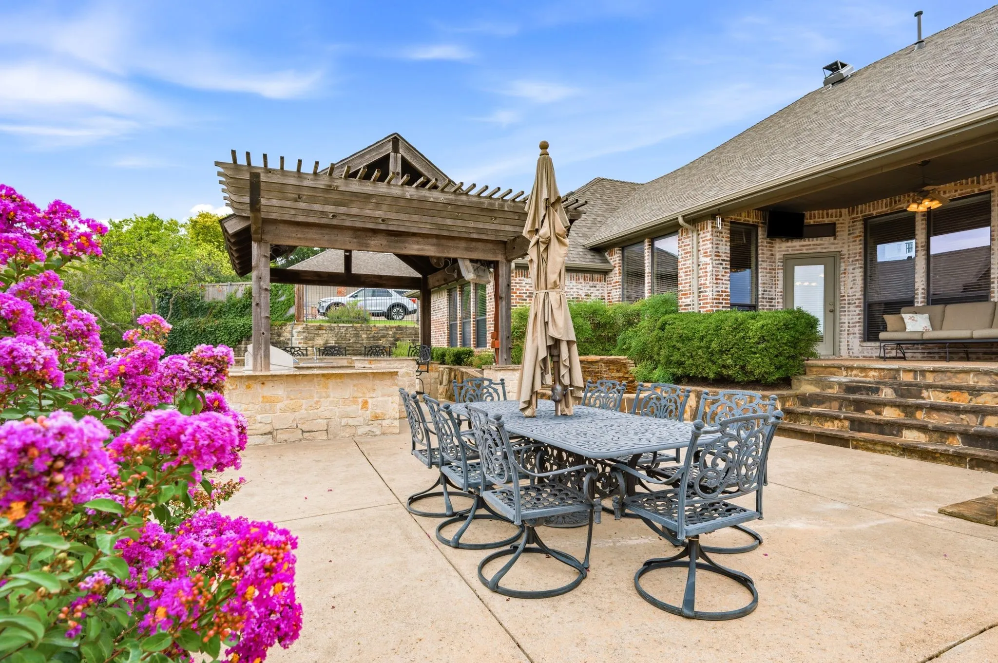 Spacious concrete patio featuring a substantial pergola, stone-clad steps, mature flowering shrubs, and a brick exterior