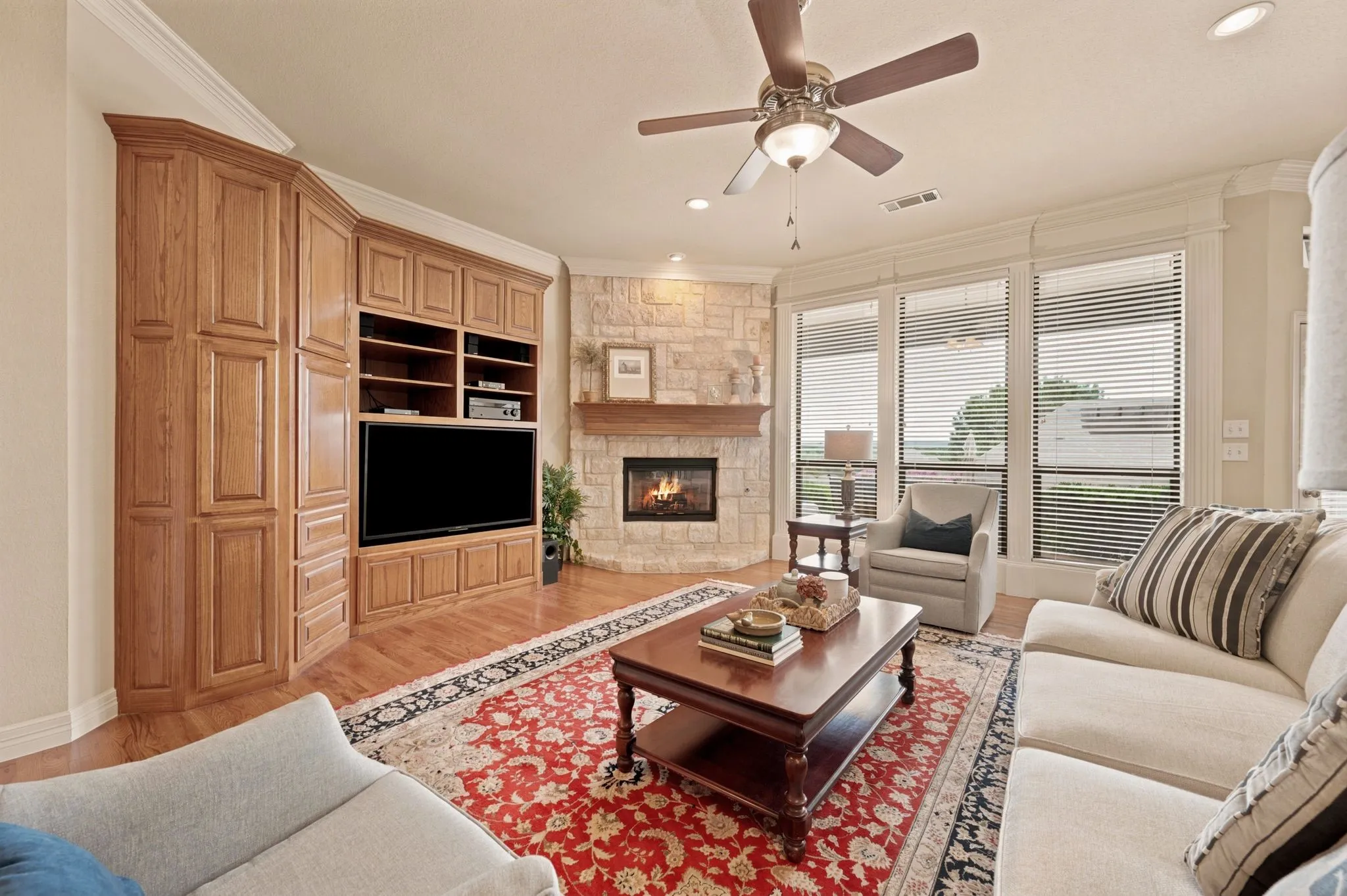 Hardwood flooring throughout featuring a corner stone fireplace with a wood mantle, built-in media cabinetry, and multiple windows with horizontal blinds
